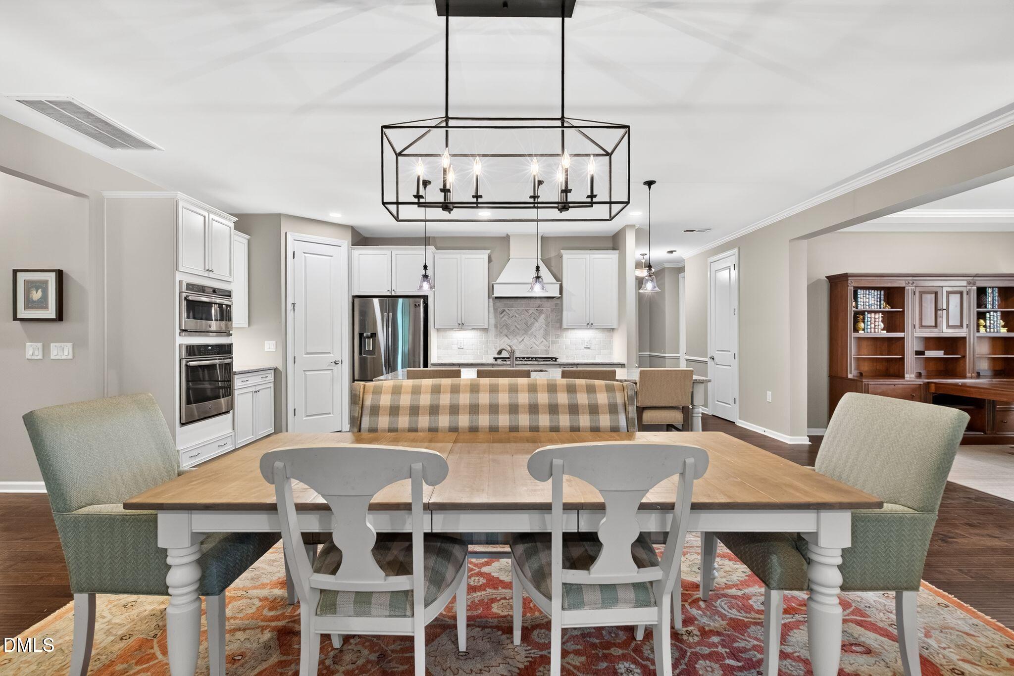 Dining room with a large wood table and black chandelier, opening to a white-cabinet kitchen.