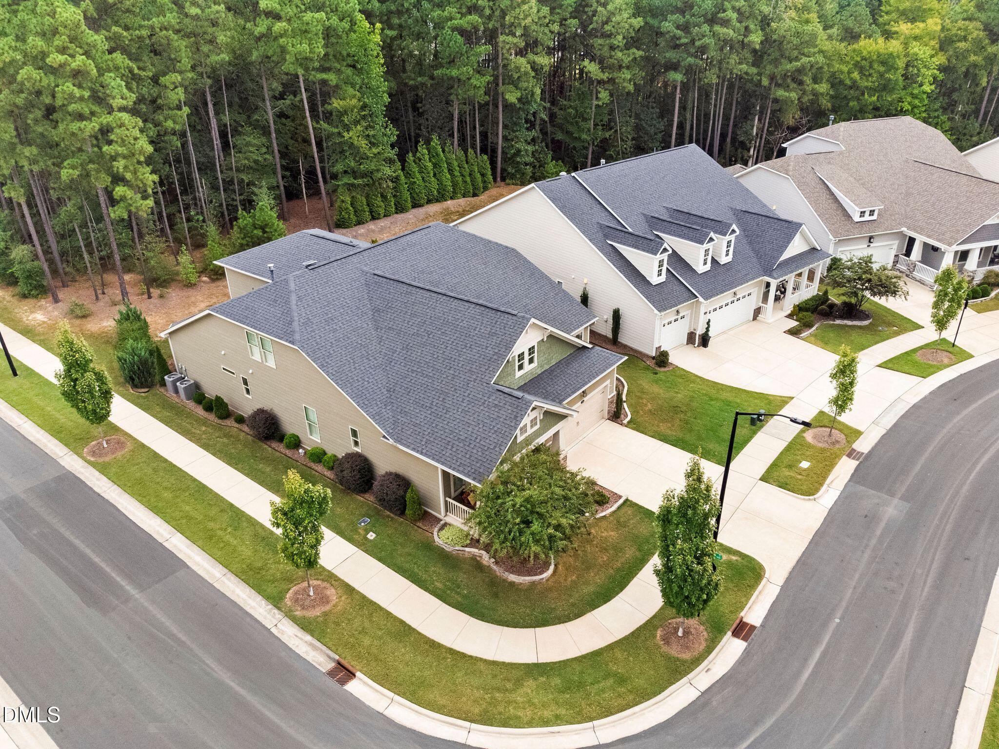 Aerial view of a beige corner-lot home with a dark gray roof and wooded backdrop.