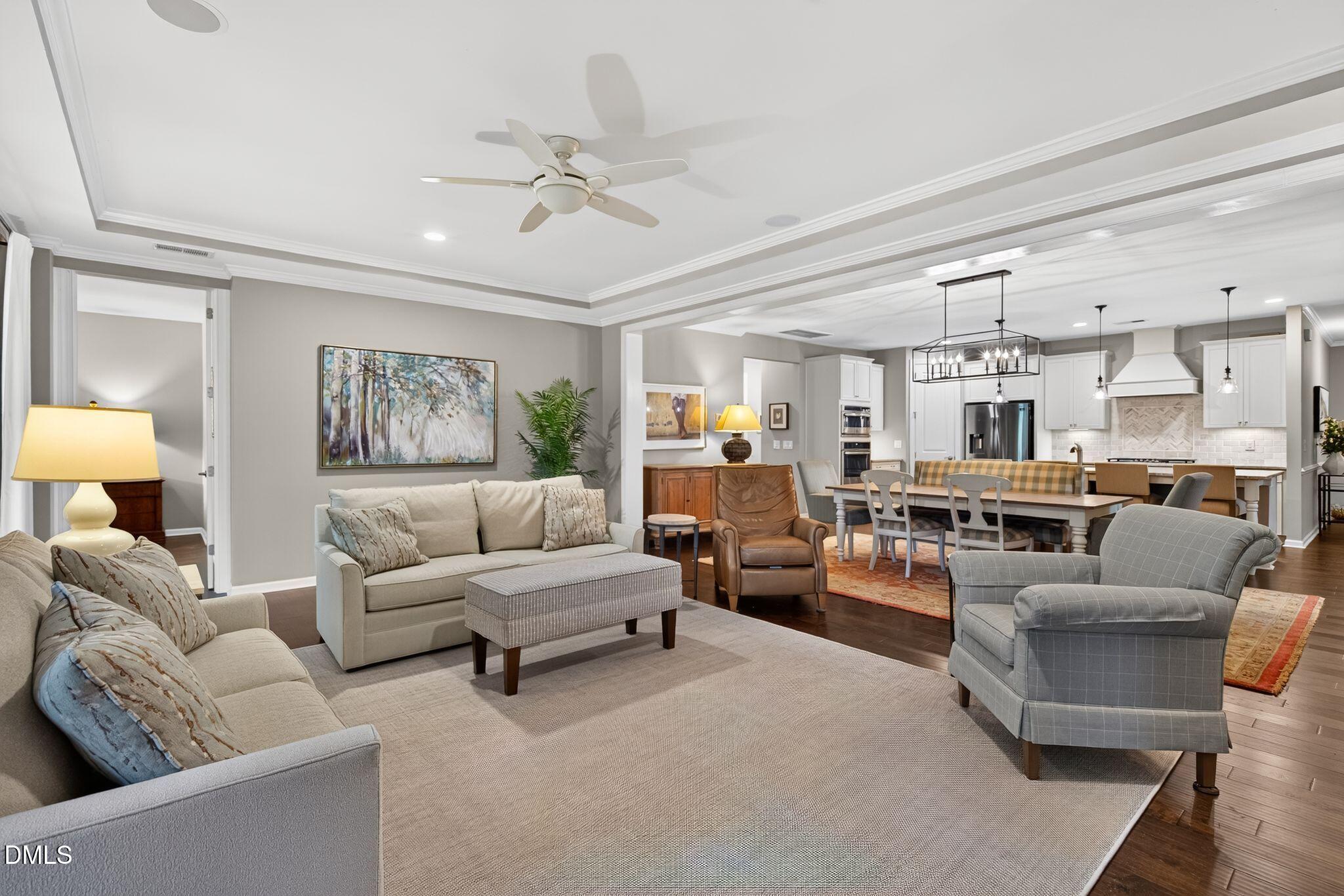 Open-plan living room and kitchen with neutral seating, tray ceiling, hardwood floors, and pendant-lit dining area.