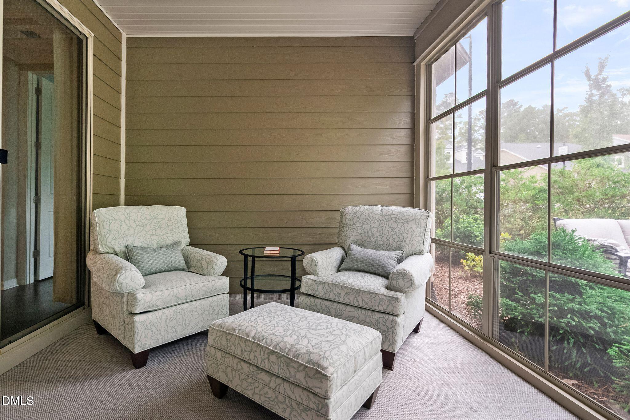 Furnished sunroom with patterned armchairs and ottoman beside large windows overlooking greenery.