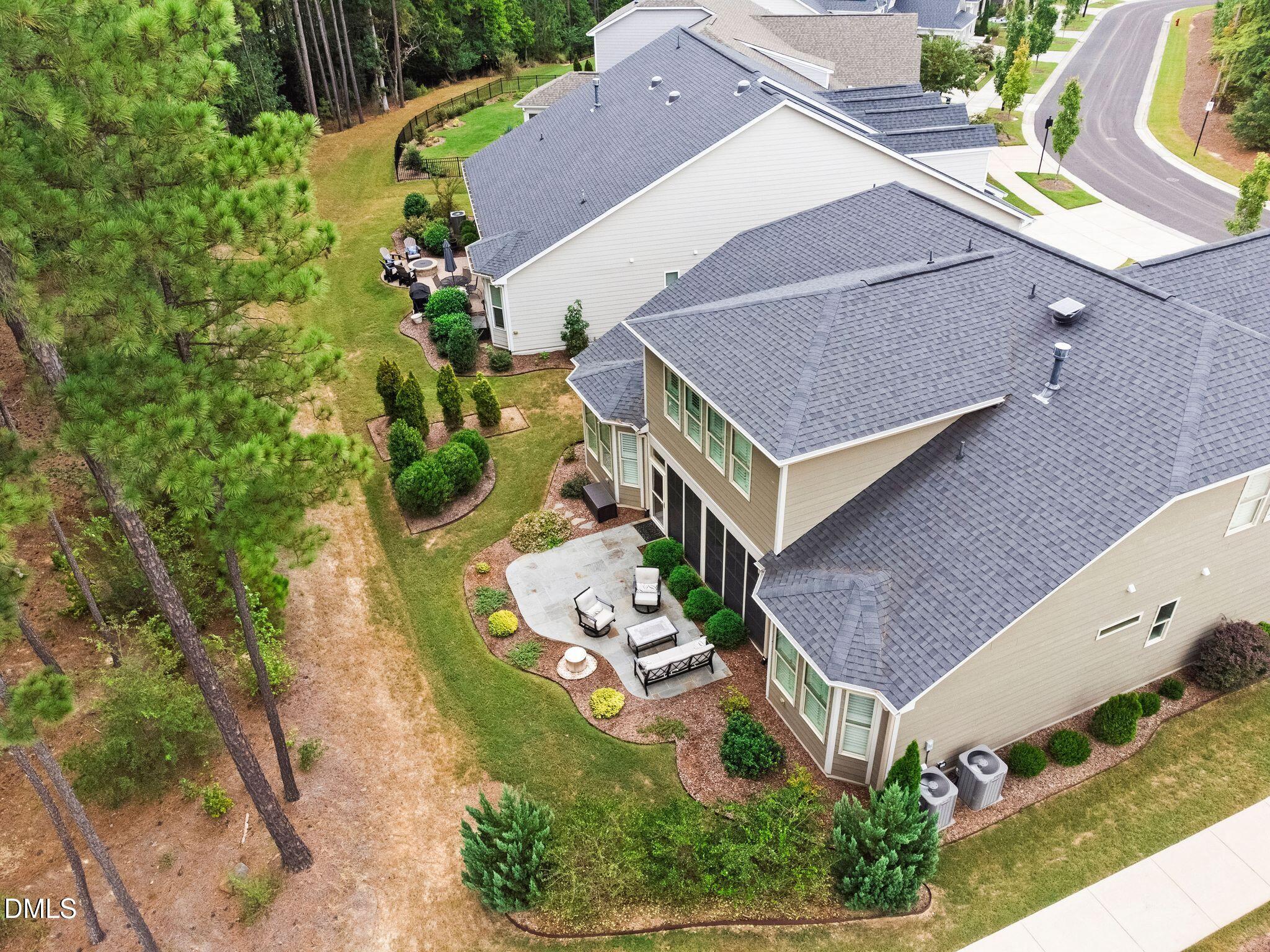 Aerial view of a beige house with a landscaped backyard, patio seating, screened porch, and surrounding pine trees.