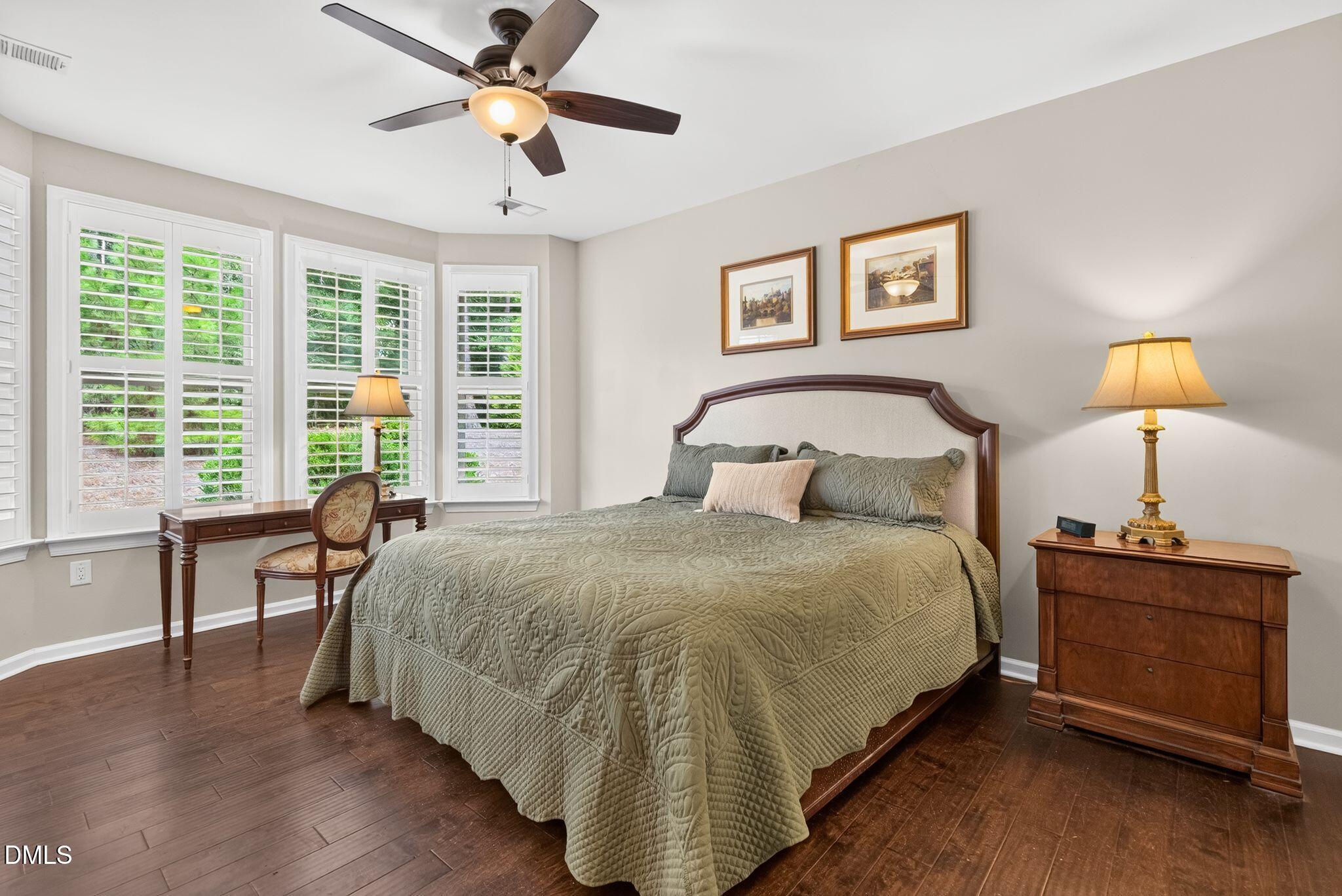 Bedroom with a sage-green quilted bed, plantation shutters, and dark wood flooring.