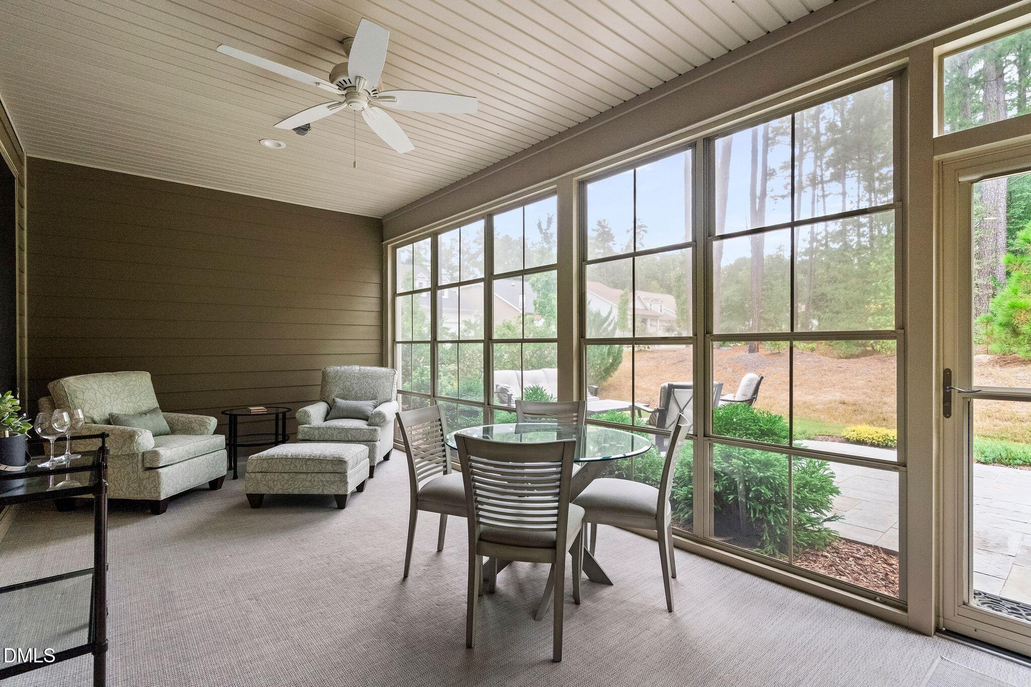Furnished sunroom with a glass dining table, upholstered chairs, and large windows overlooking the patio and trees.