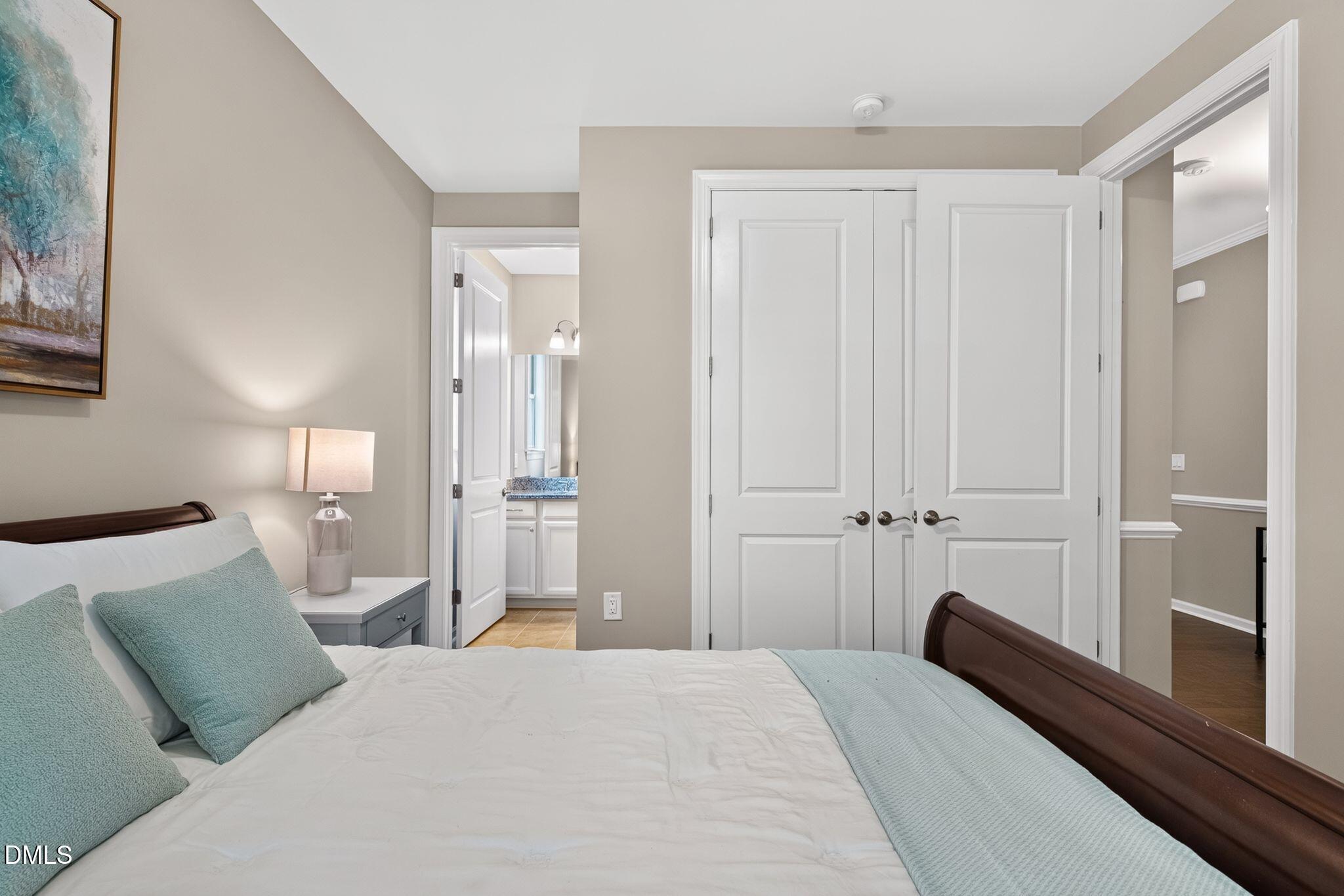 Staged bedroom with a white bed, pale blue-green accents, white closet doors, and a glimpse of an adjoining bathroom.