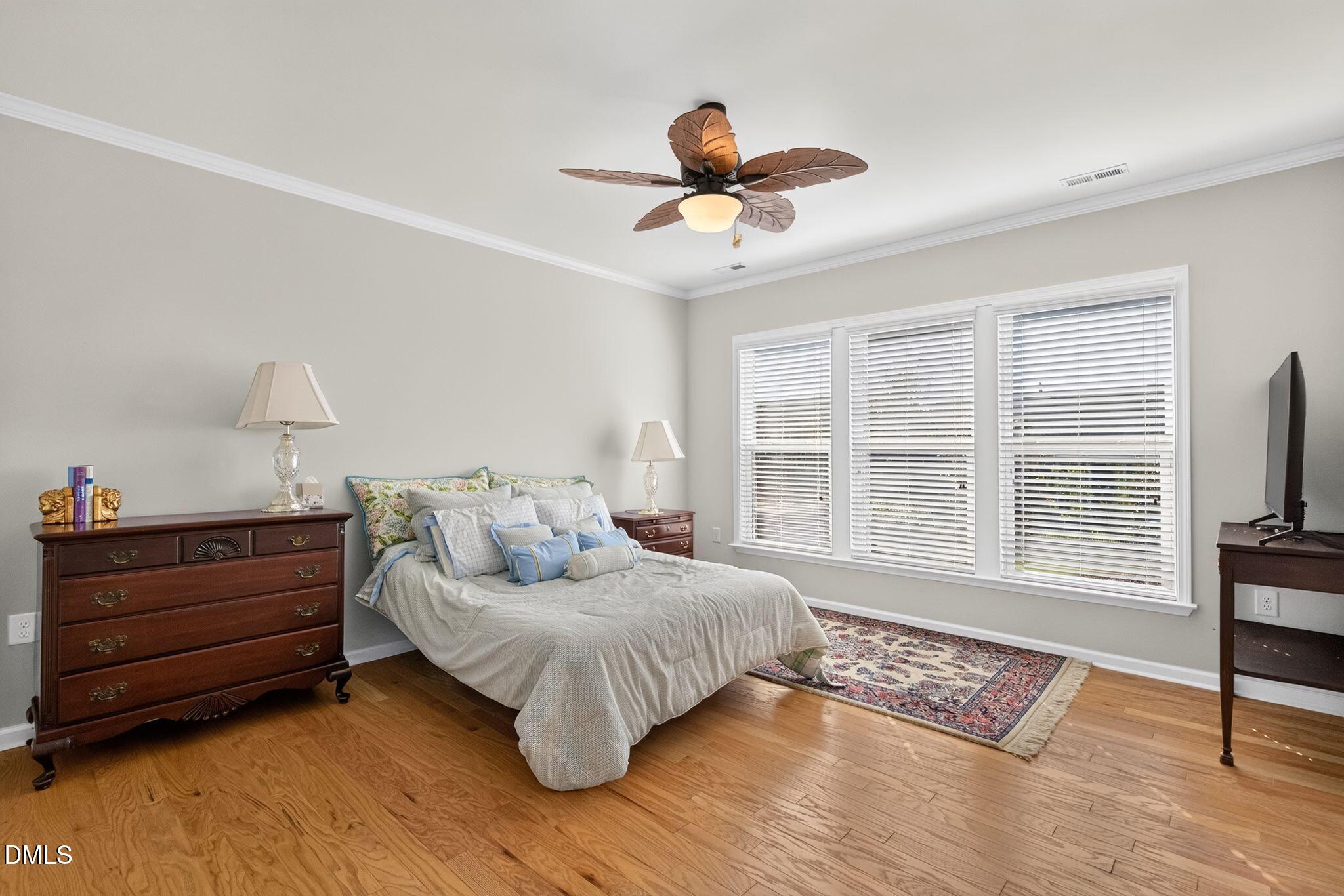 Furnished bedroom with a white-and-blue bed, hardwood floors, and three wide windows with blinds.