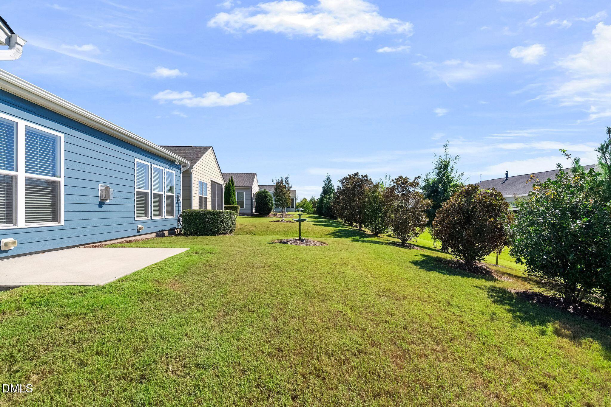 Residential side yard with a broad lawn, concrete patio, and trimmed shrubs beside blue-sided homes.