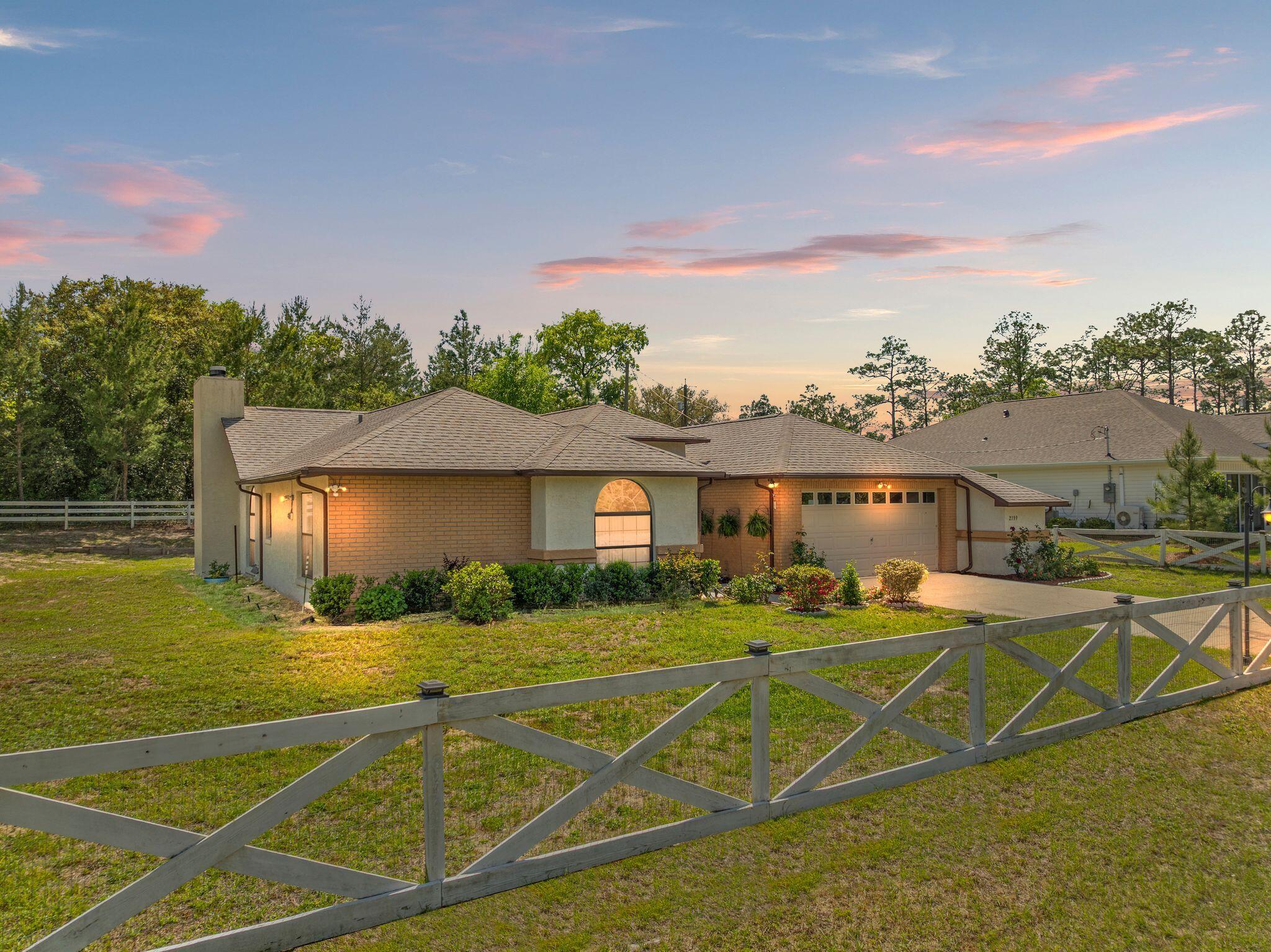 Single-story brick home with attached garage behind a cross-braced fence at dusk.
