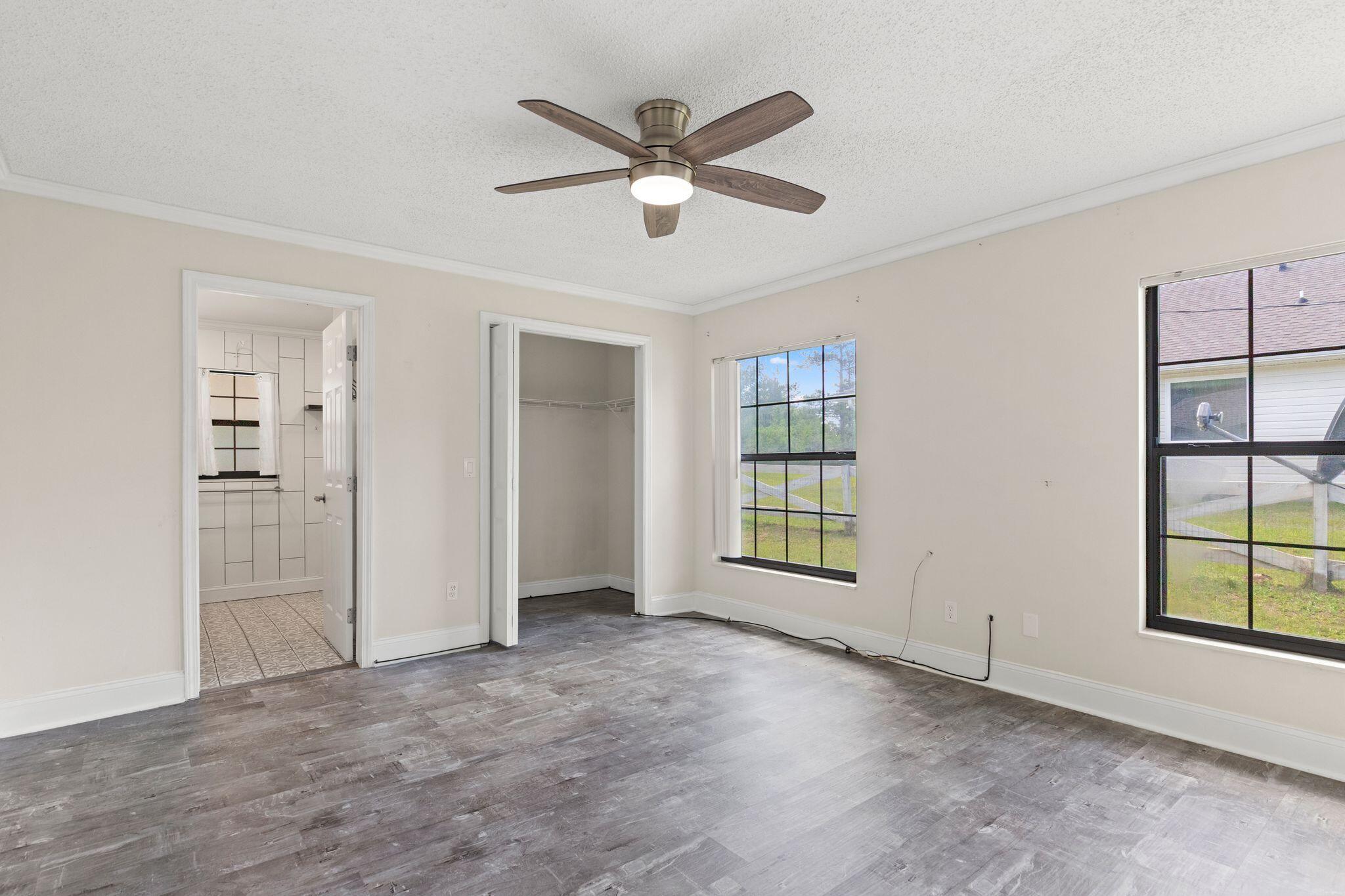 Empty bedroom with gray wood-look flooring, a ceiling fan, an open closet, and two large windows.