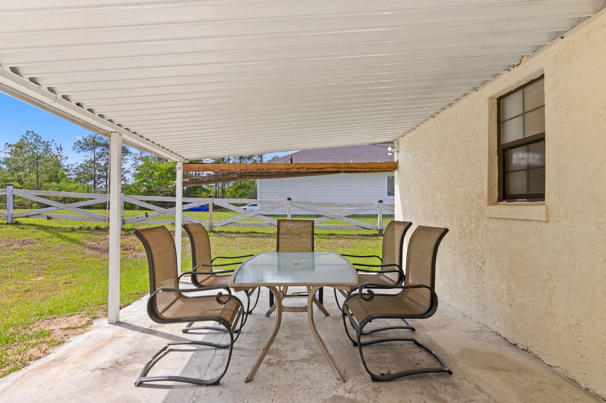 Covered patio with a glass-top dining table and chairs beside a fenced grassy yard.