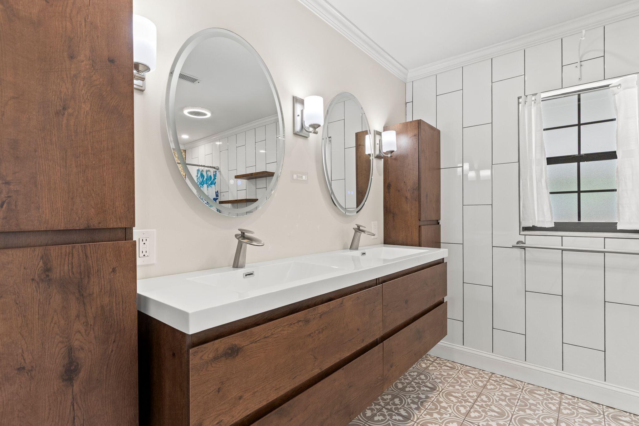 Bathroom with a dark wood double vanity, round mirrors, white tiled walls, and patterned floor.