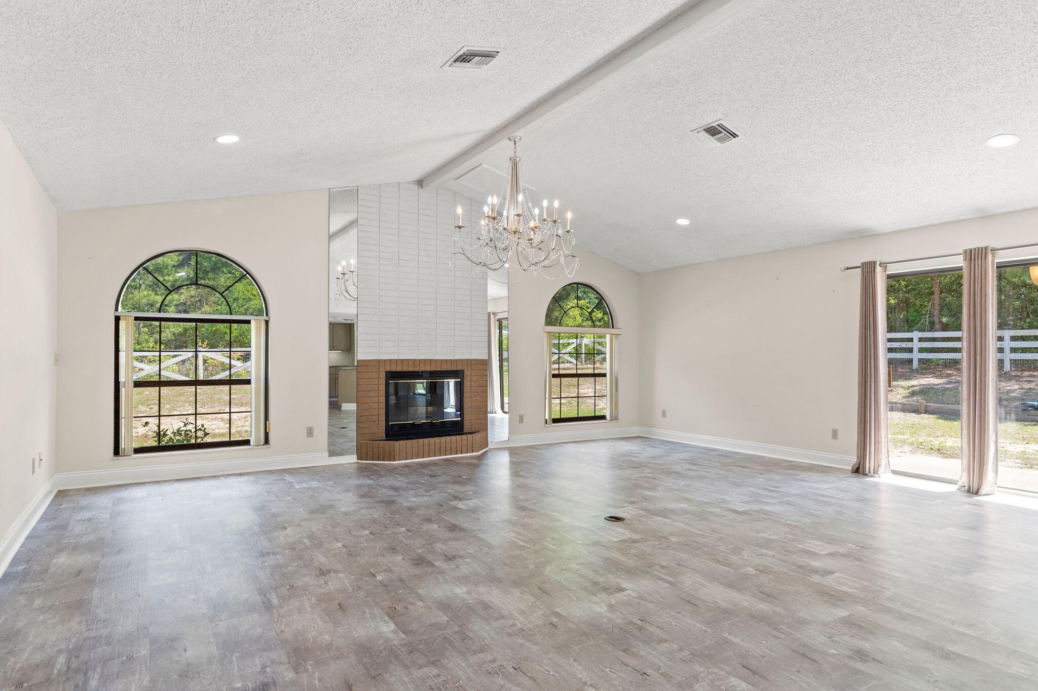 Vacant living room with a brick fireplace, chandelier, and arched windows overlooking a fenced yard.