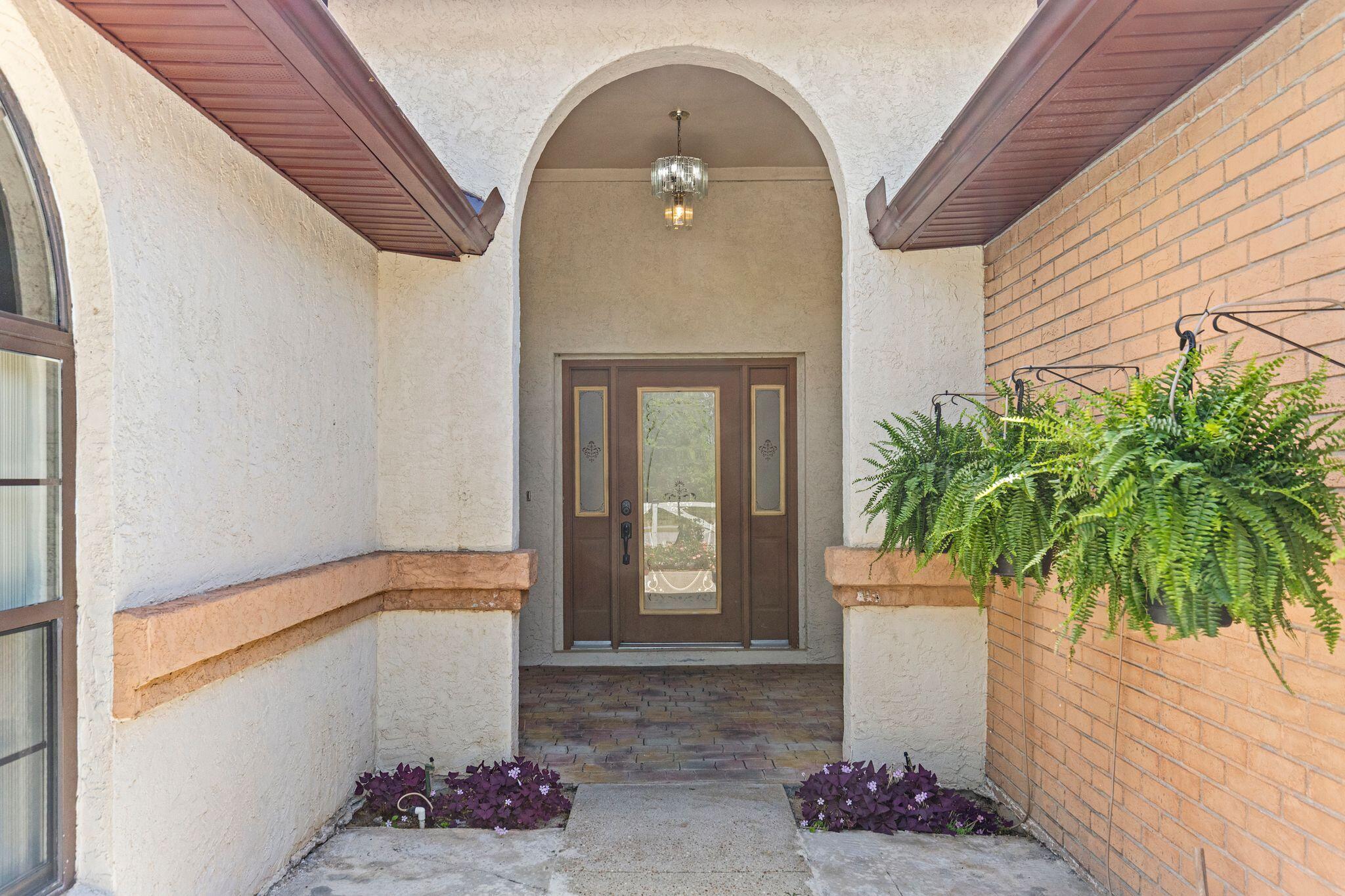 Recessed front entry with arched stucco surround, glass-paneled double doors, brick paving, and hanging fern.