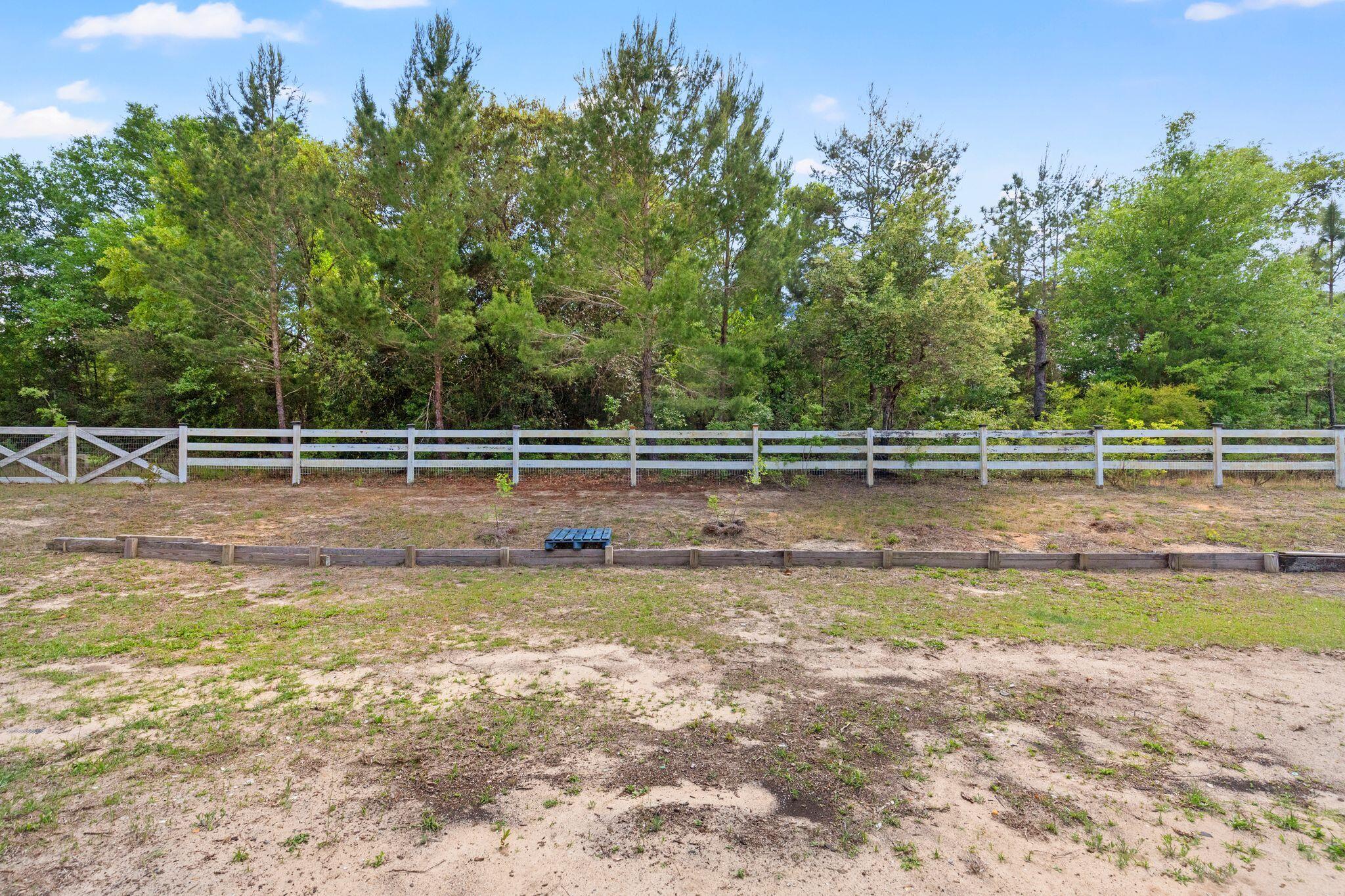 Sandy lot enclosed by a white rail fence, with mature trees and woods behind it.