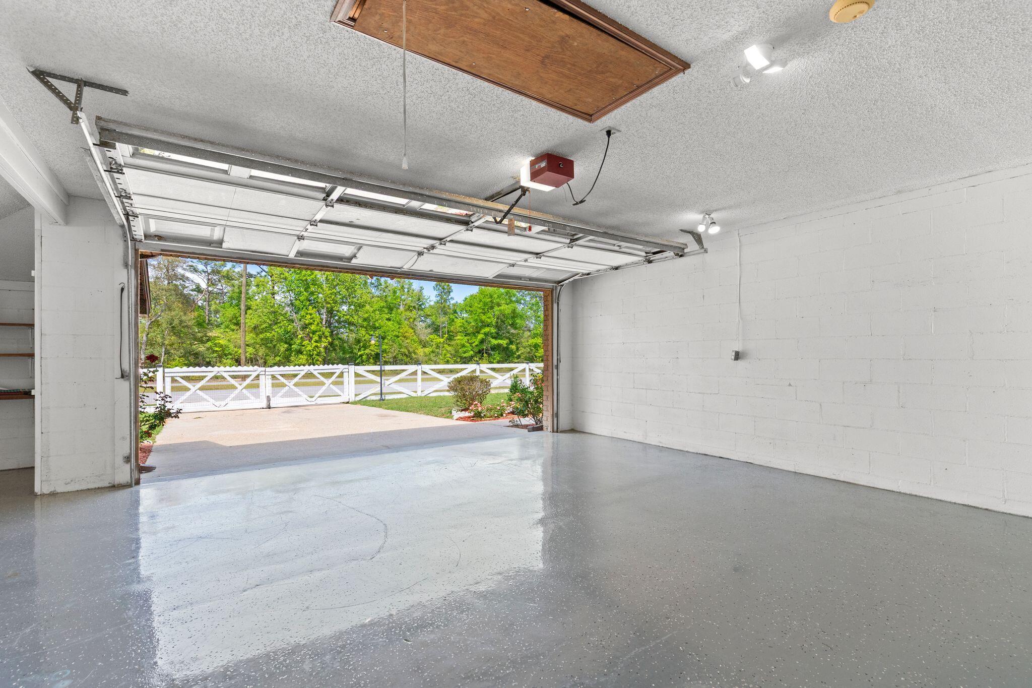 Empty garage with a glossy gray floor and open door facing a white fence and trees.