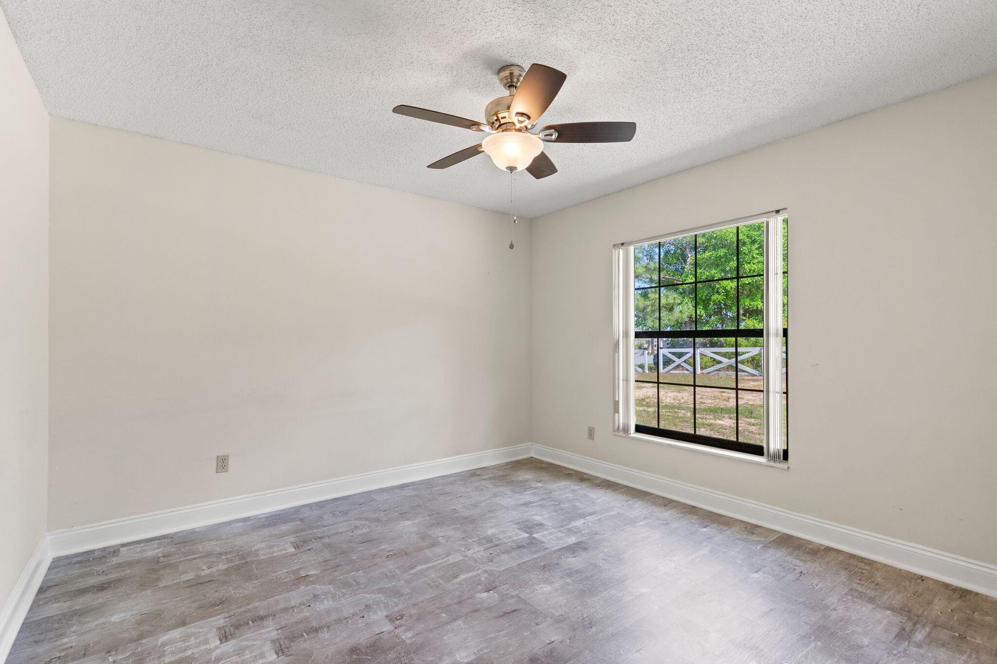 Empty room with gray wood-look flooring, ceiling fan, and a large window overlooking trees and a white fence.