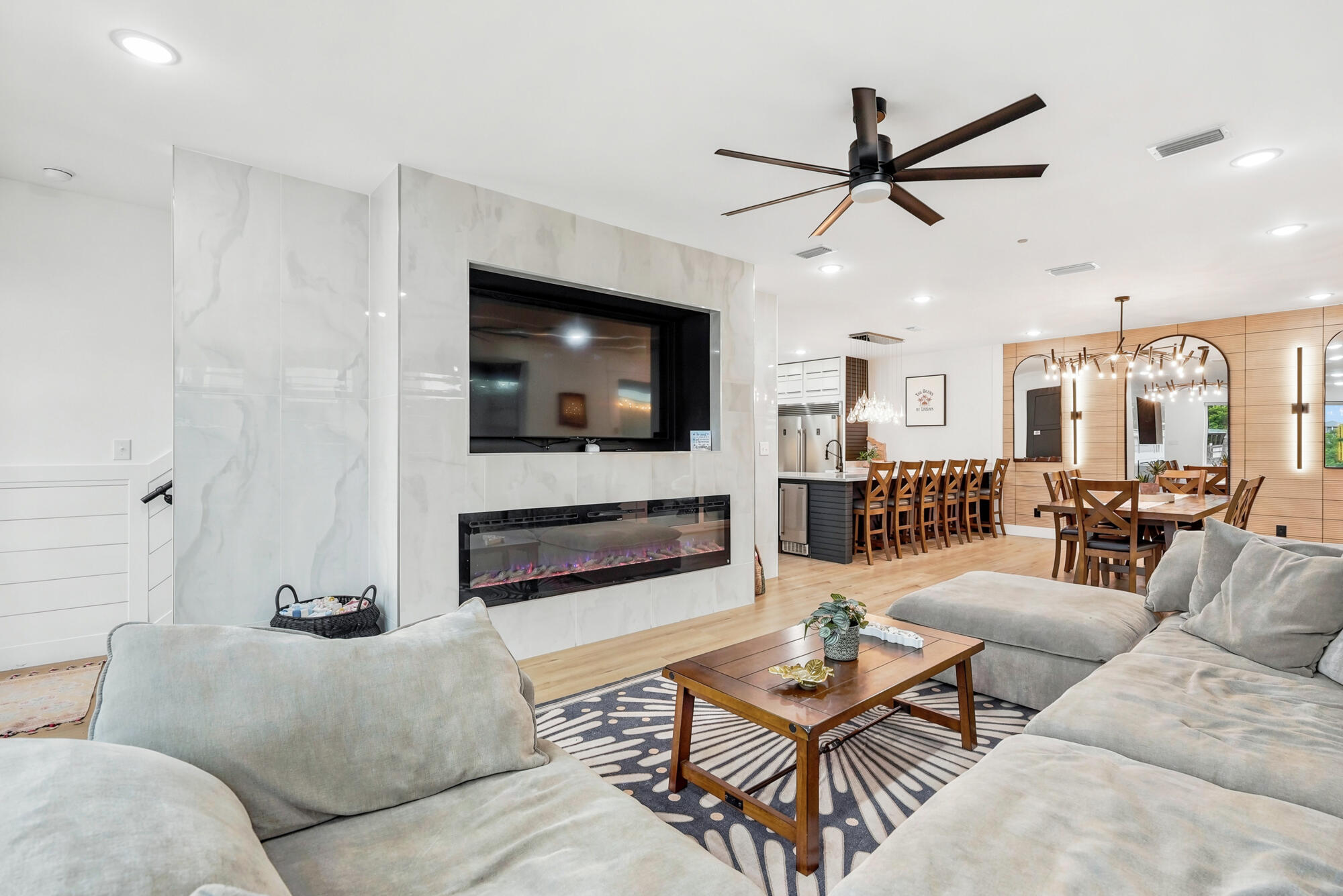 Open-plan living and dining room with a marble-look fireplace wall, pale sectional sofas, and a wood dining set.