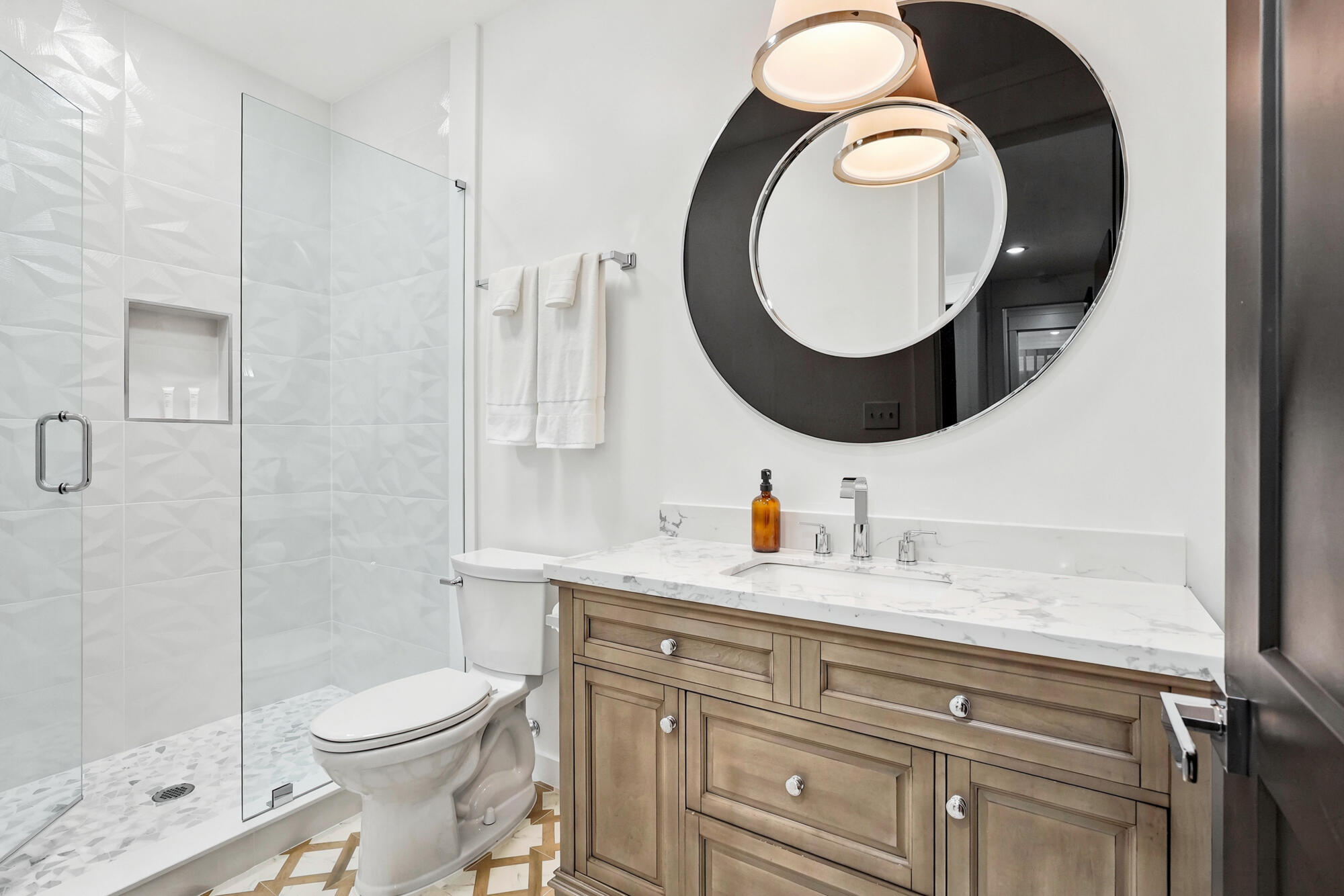 Modern bathroom with glass walk-in shower, geometric tile, wood vanity, and oversized round mirror.