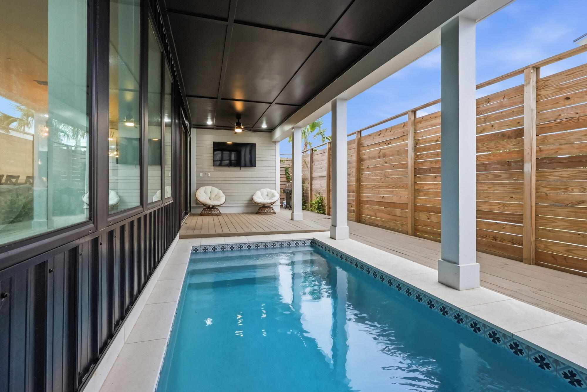 Outdoor pool beside a covered patio with lounge chairs and a tall wooden privacy fence.