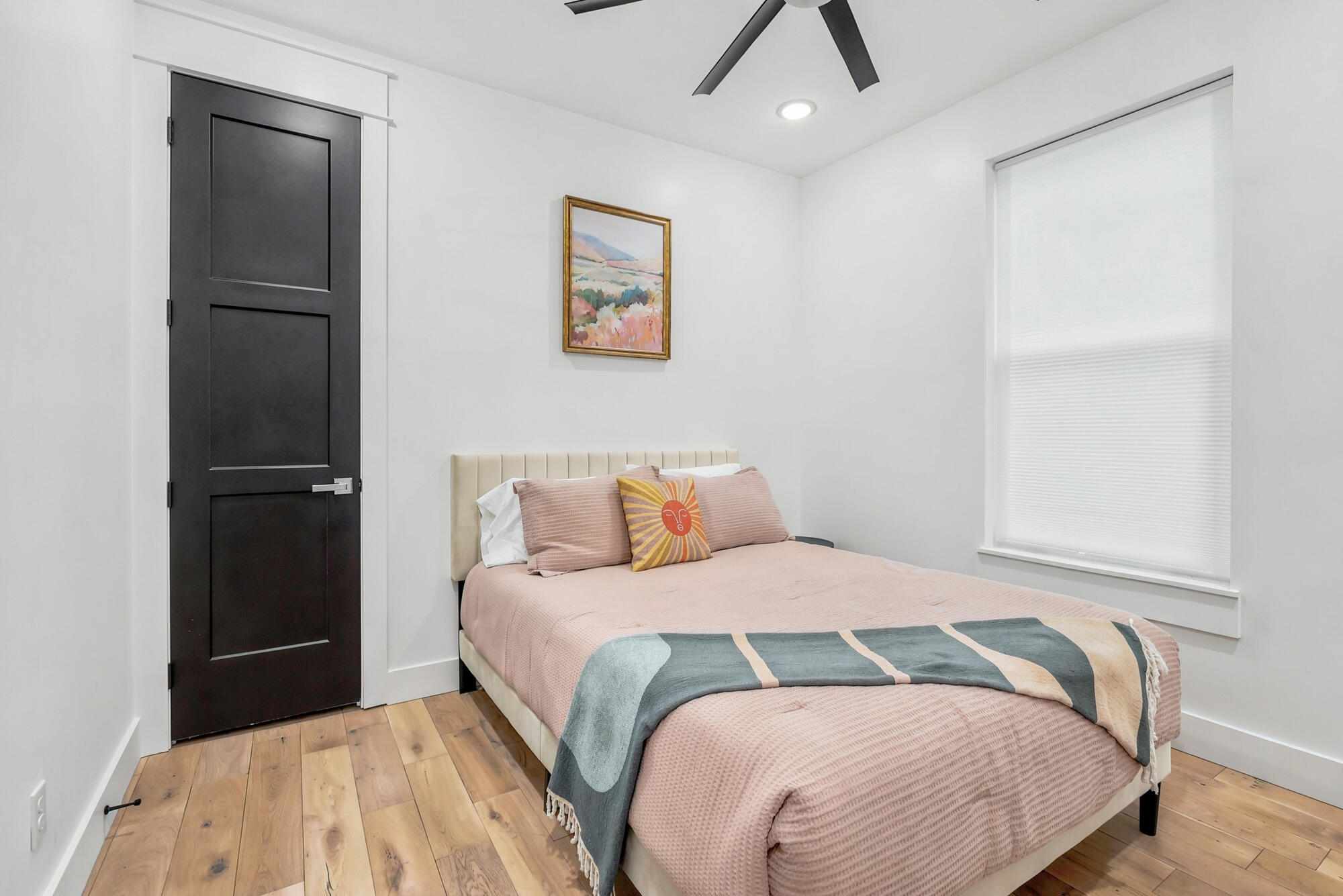 Staged bedroom with a blush-covered bed, patterned throw, black paneled door, and light wood flooring.
