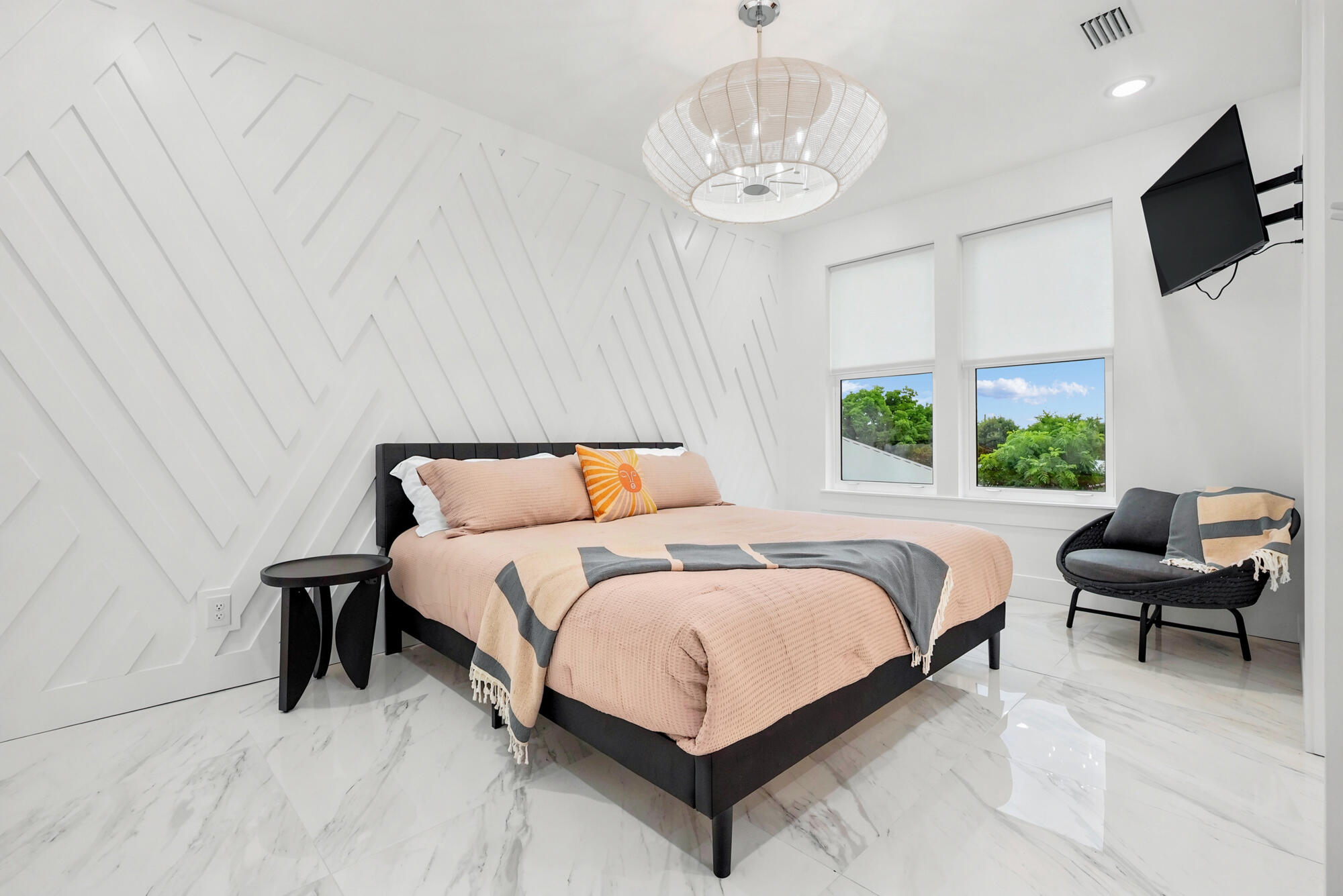 Staged bedroom with blush-pink bedding, a geometric white accent wall, marble-look floors, and a woven pendant light.