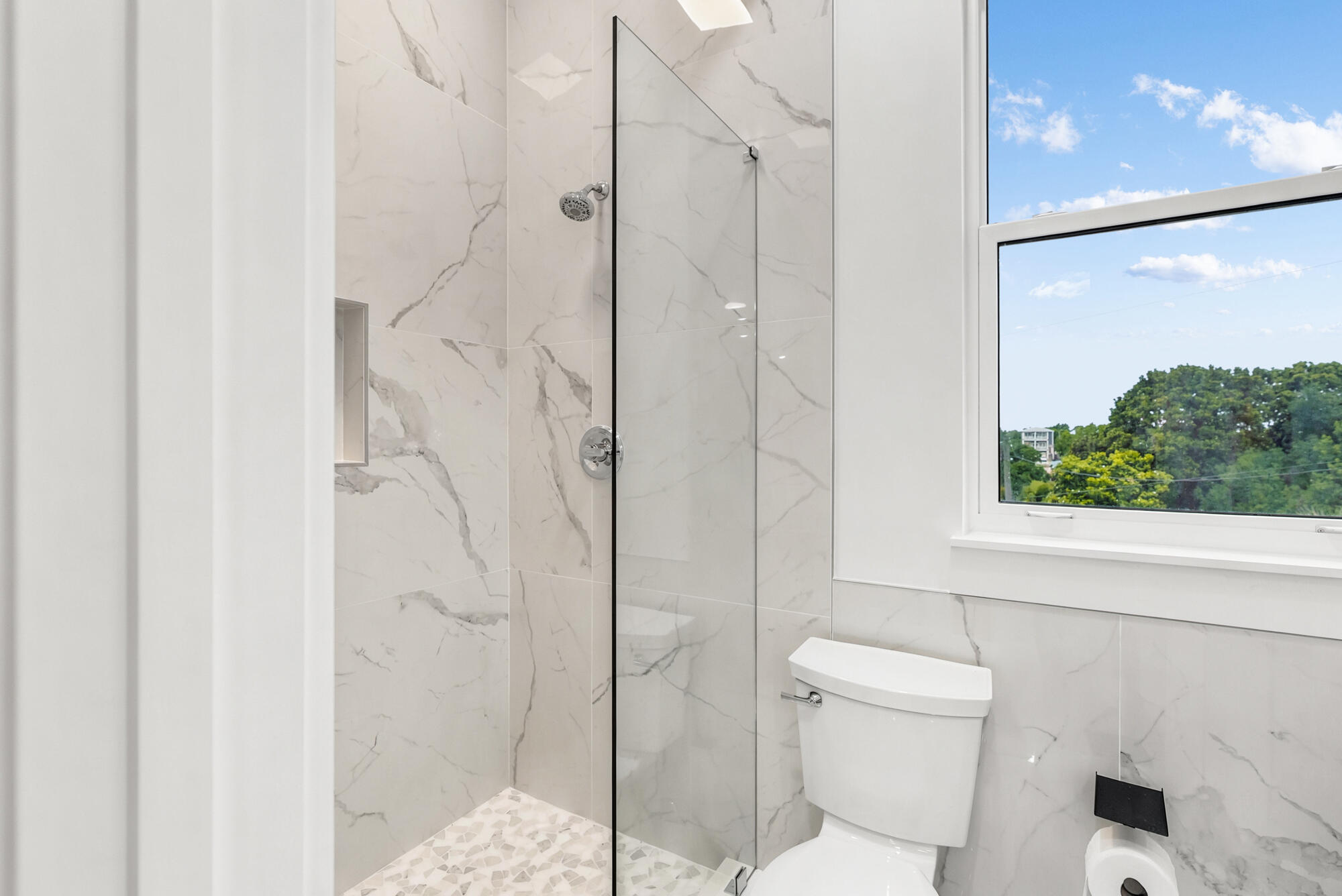 Modern marble-look bathroom with a glass walk-in shower and a window overlooking trees.