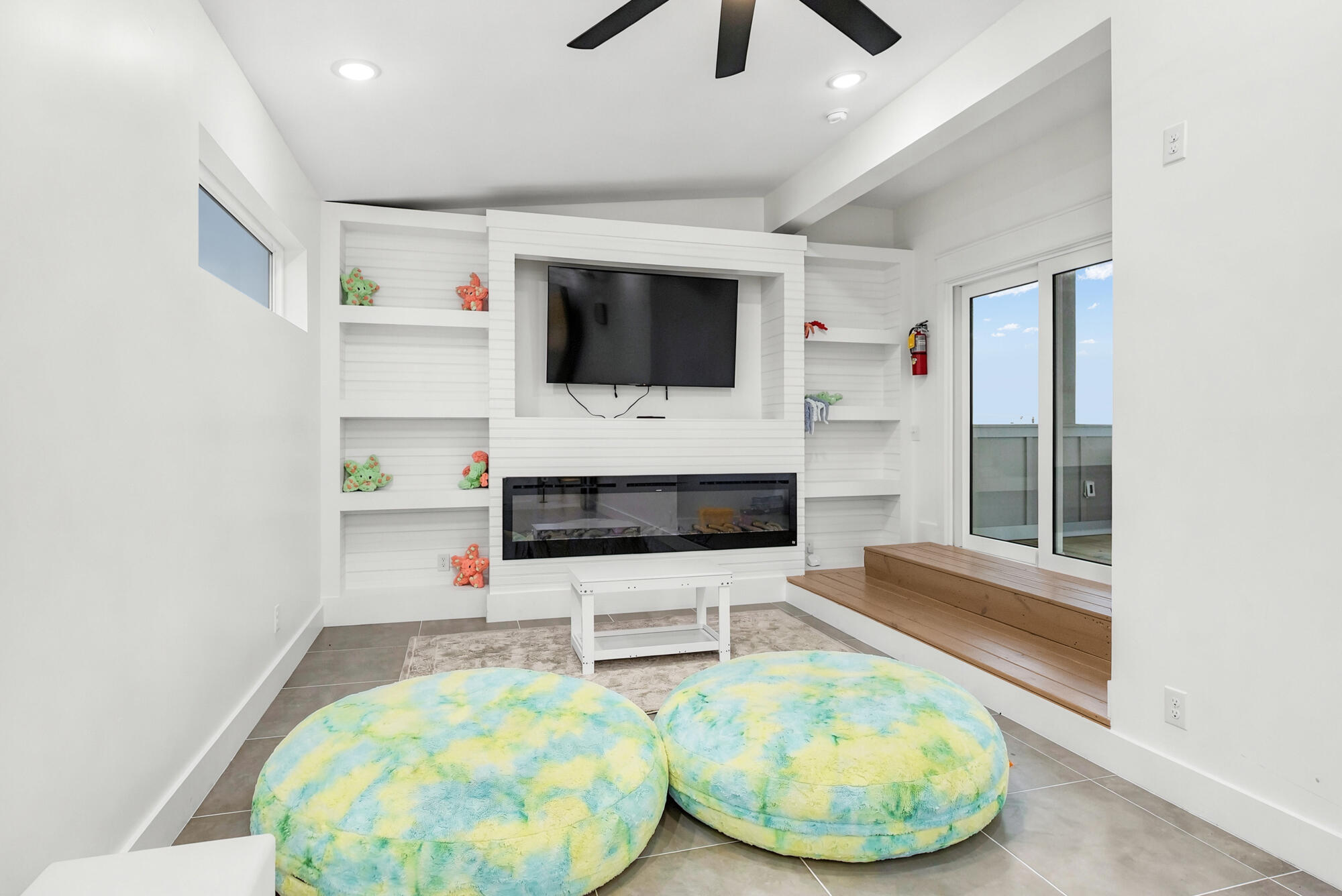 Living room with a TV above a linear fireplace, built-in shelves, colorful floor cushions, and a wood bench.