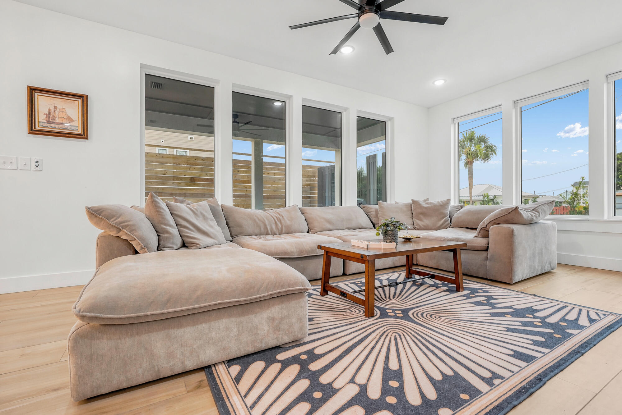 Furnished living room with a beige sectional, patterned rug, and tall windows overlooking a palm tree.