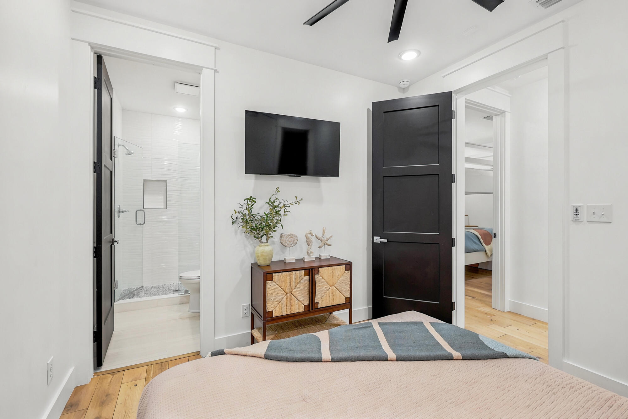 Staged bedroom with ensuite bathroom, wall-mounted TV, dark doors, and light wood flooring.