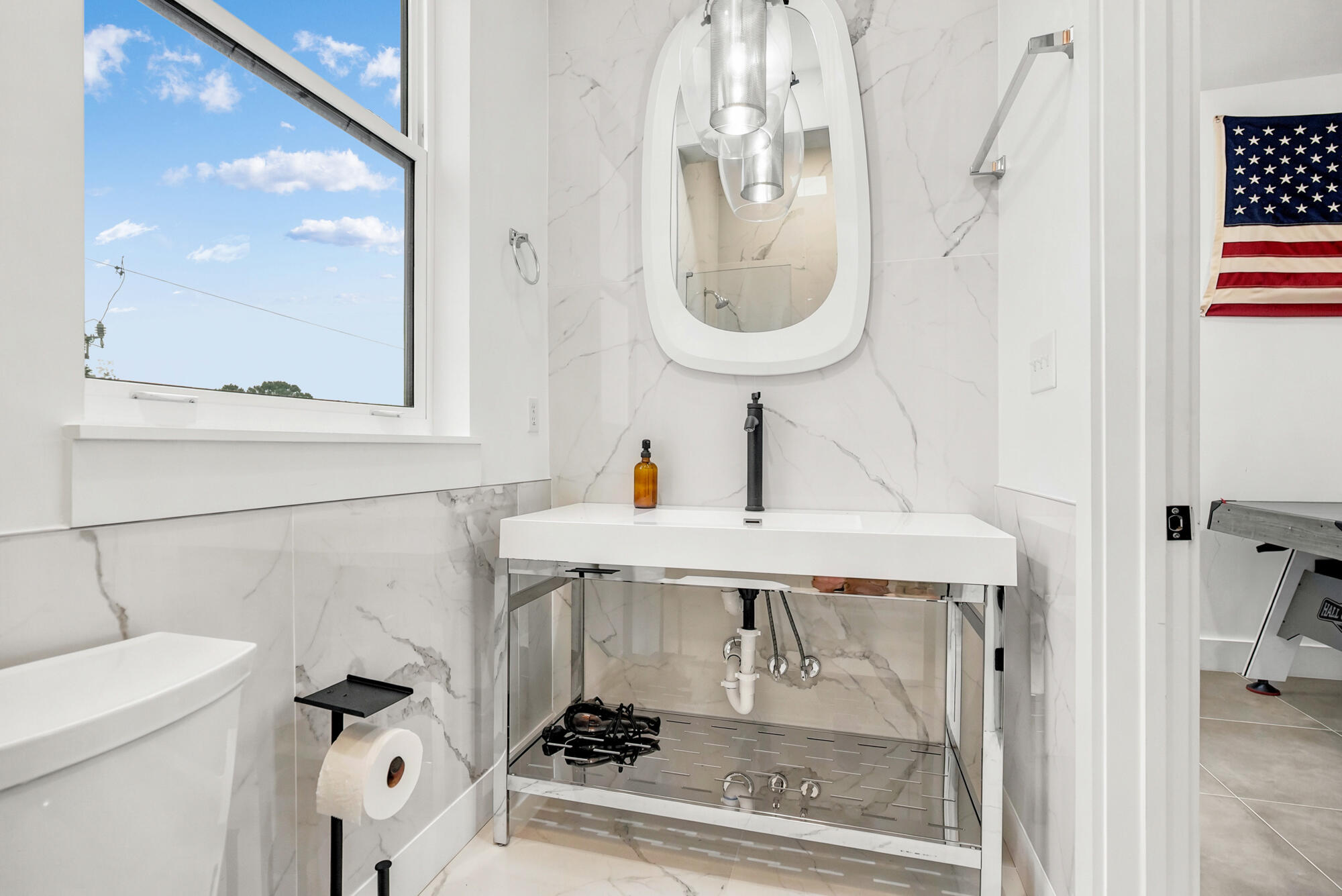 Modern marble-look bathroom with a white vanity, black faucet, rounded mirror, and large window.