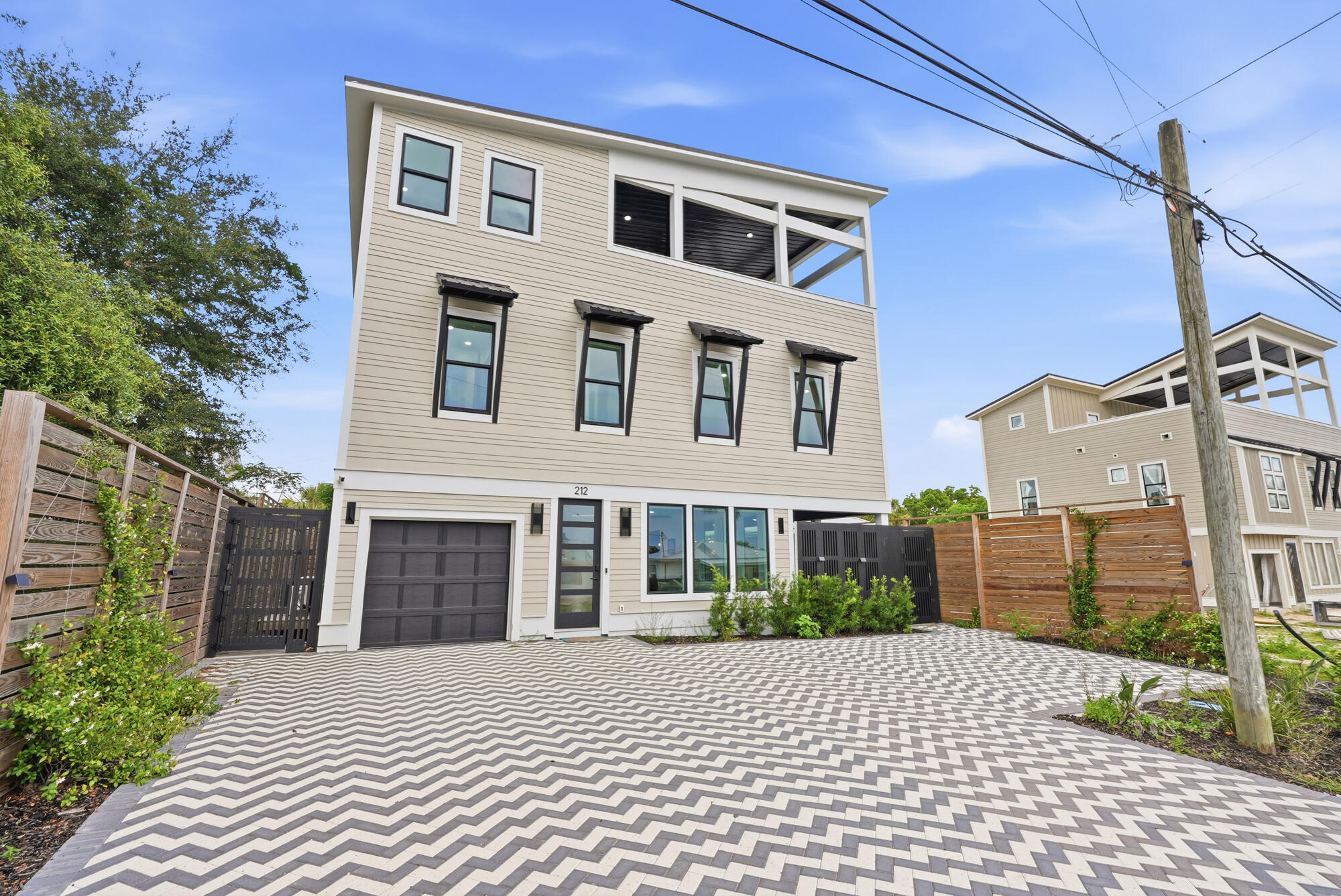 Three-story beige home with a covered upper terrace, garage, and zigzag-patterned paver driveway.