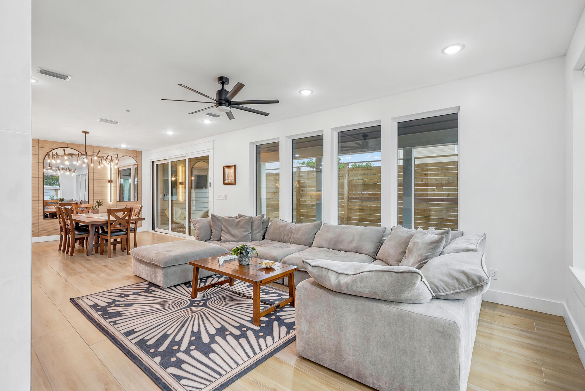 Open-plan living and dining room with a large gray sectional, patterned rug, and tall windows facing a wood fence.