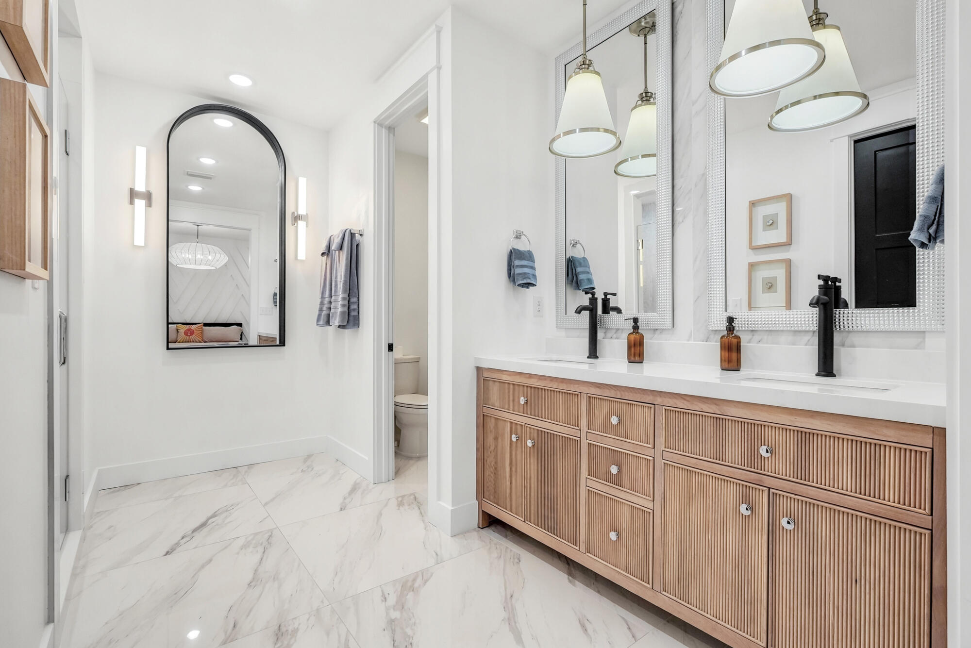 Bright bathroom with a double-sink fluted wood vanity, oversized mirrors, and marble-look tile floors.