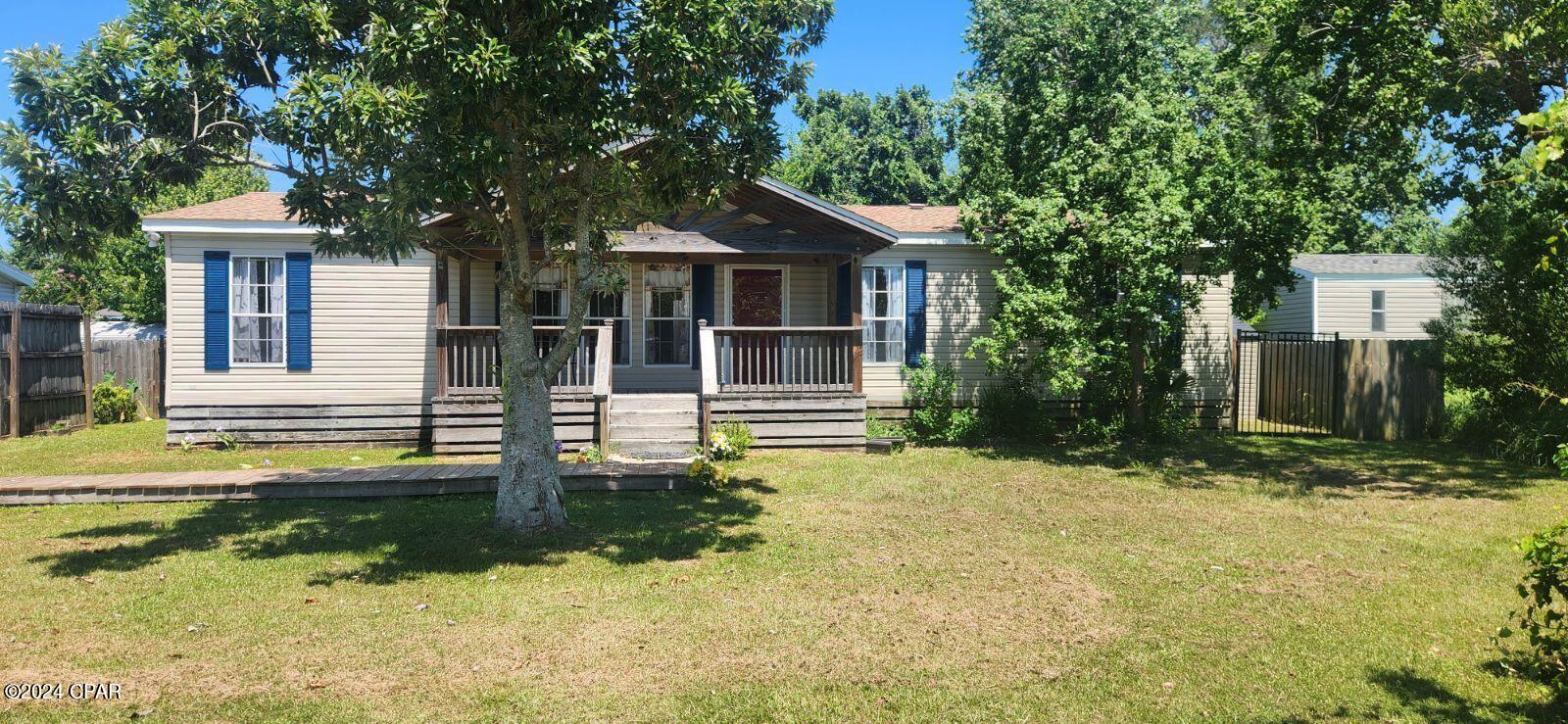 Beige home with a covered wooden porch and blue shutters beneath mature trees.