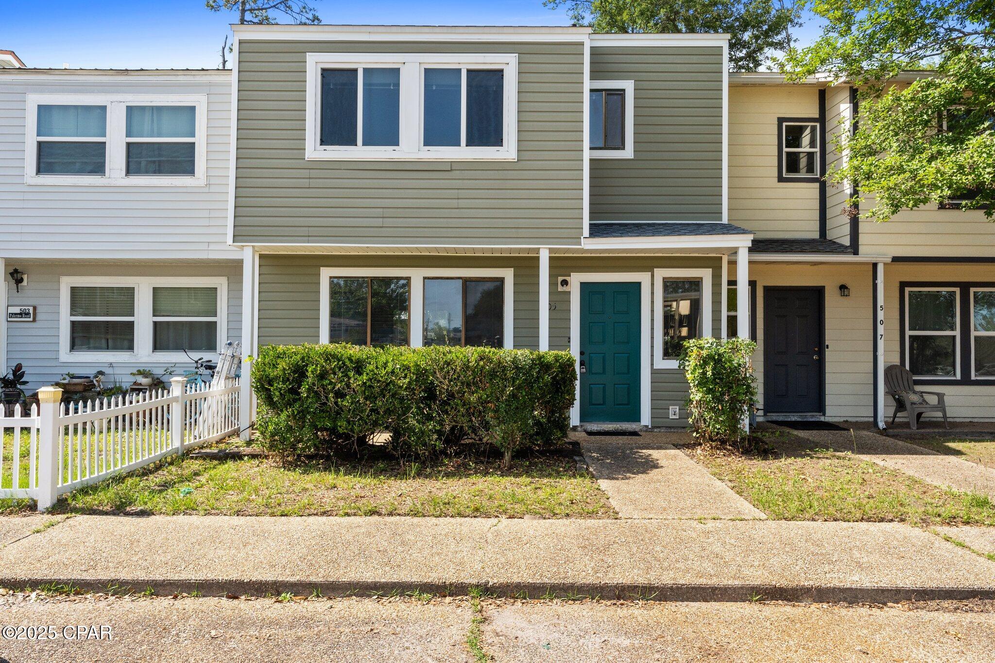 Sage-green attached home with a teal front door, white-trimmed windows, and hedges along the entry.