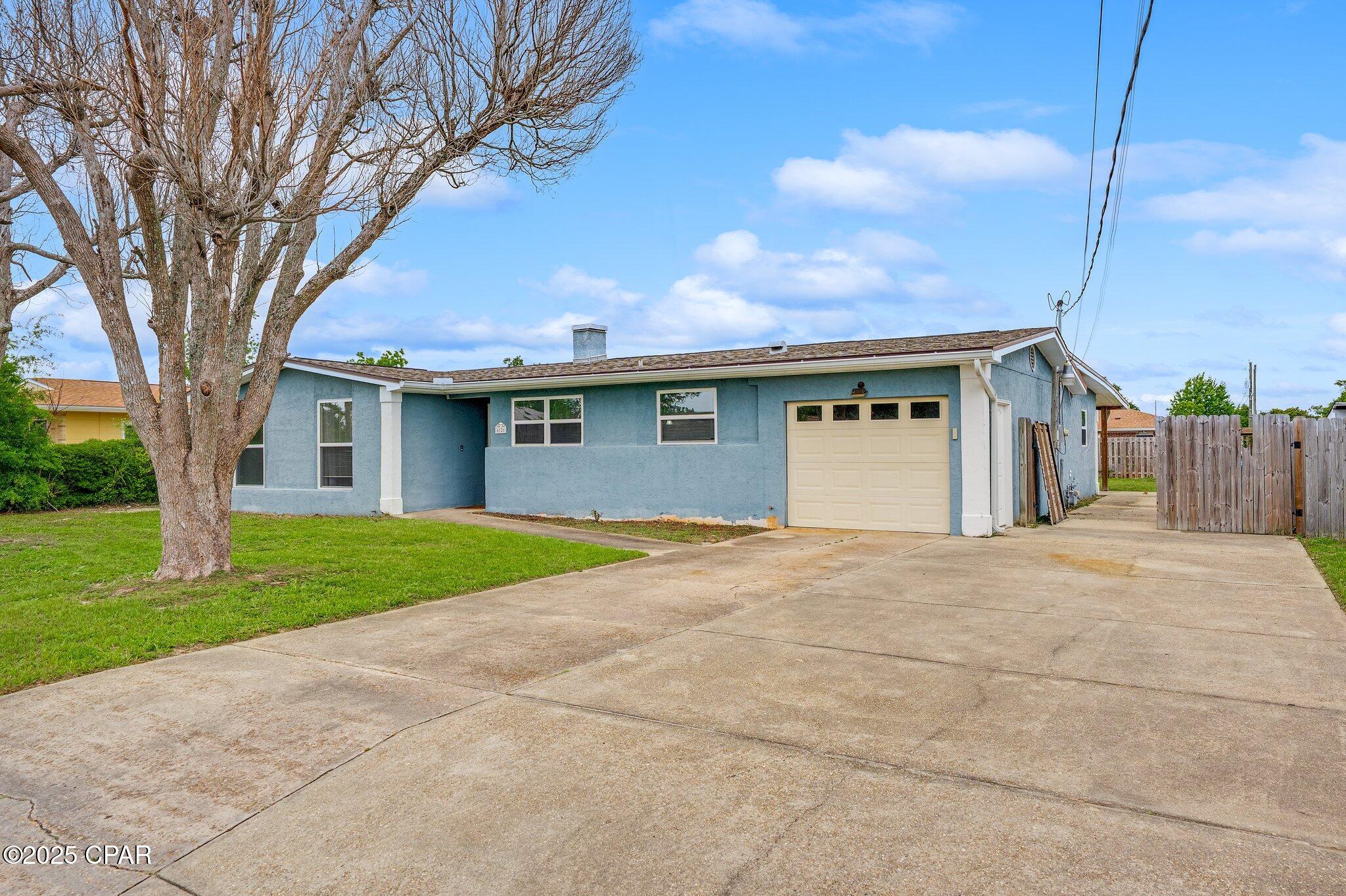 Blue single-story house with a beige garage door, wide driveway, and large leafless tree in the yard.