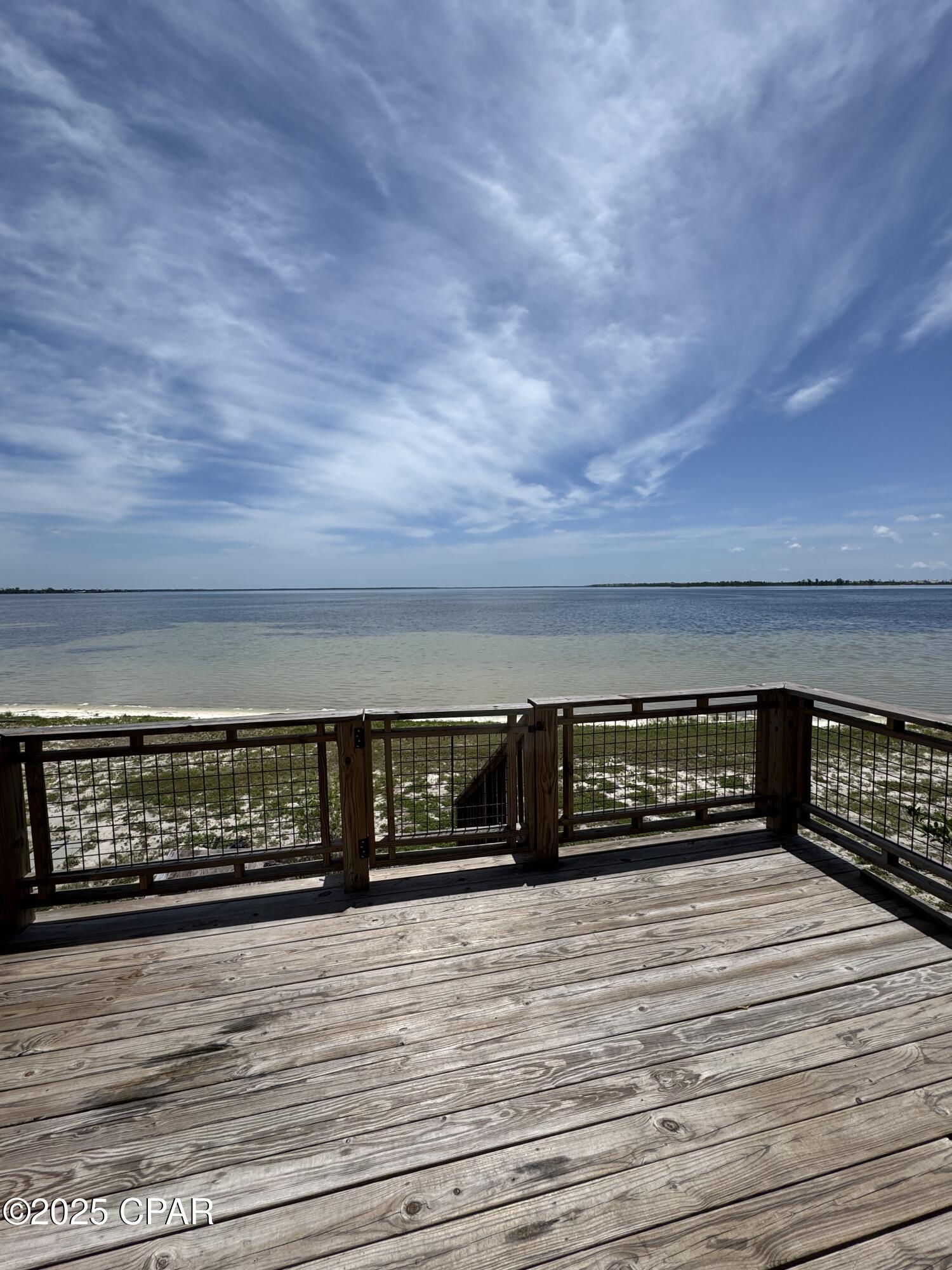 Wooden deck overlooks calm water, with a sandy shoreline and sweeping cloud-filled sky.