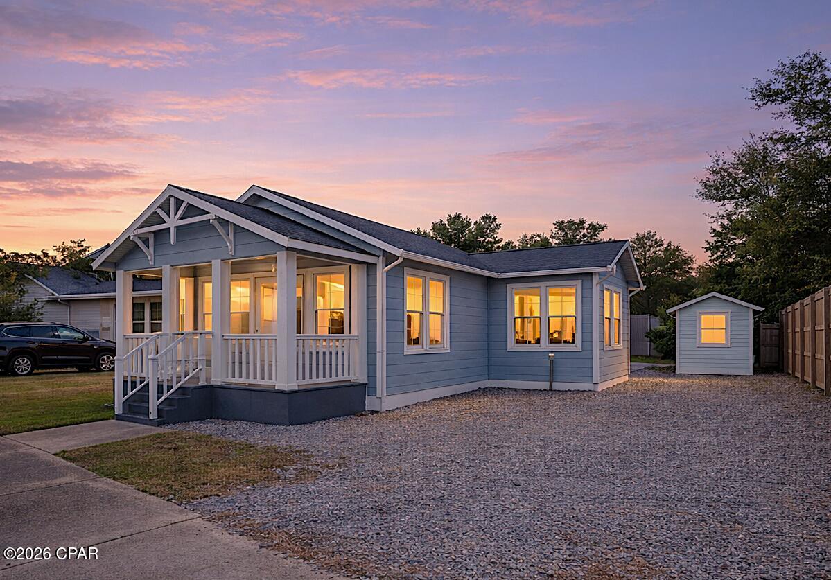 Blue single-story house with a white-railed porch and detached shed, photographed at dusk.