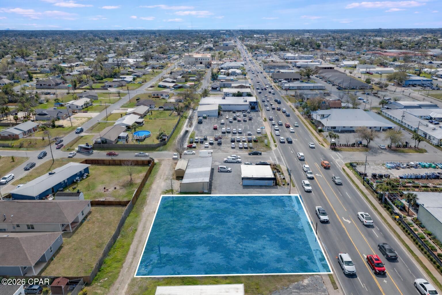 Aerial view of a blue-highlighted parcel beside a busy road in a mixed residential and commercial neighborhood.