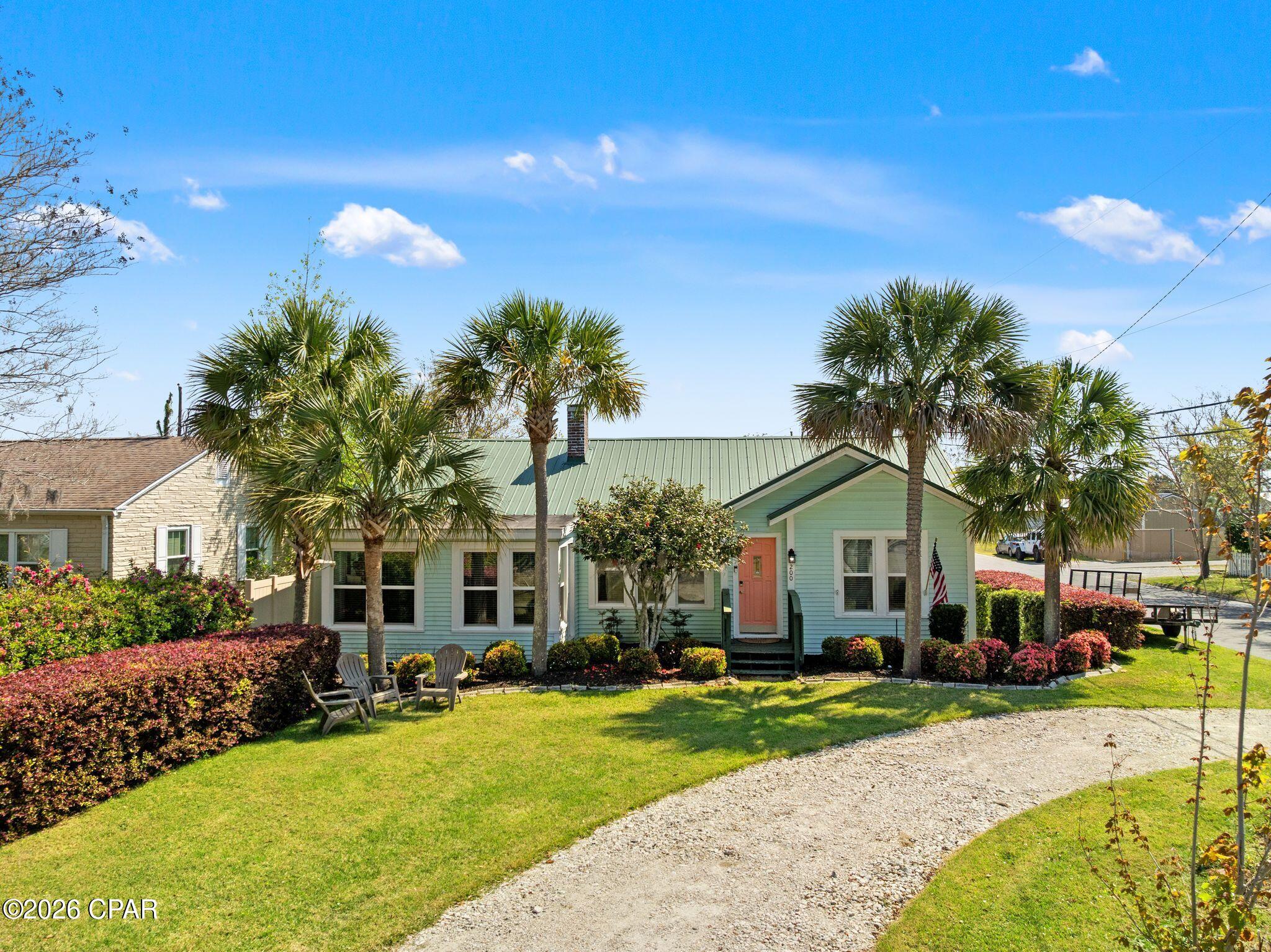 Mint-green single-story home with a green metal roof, coral door, palm trees, and gravel driveway.