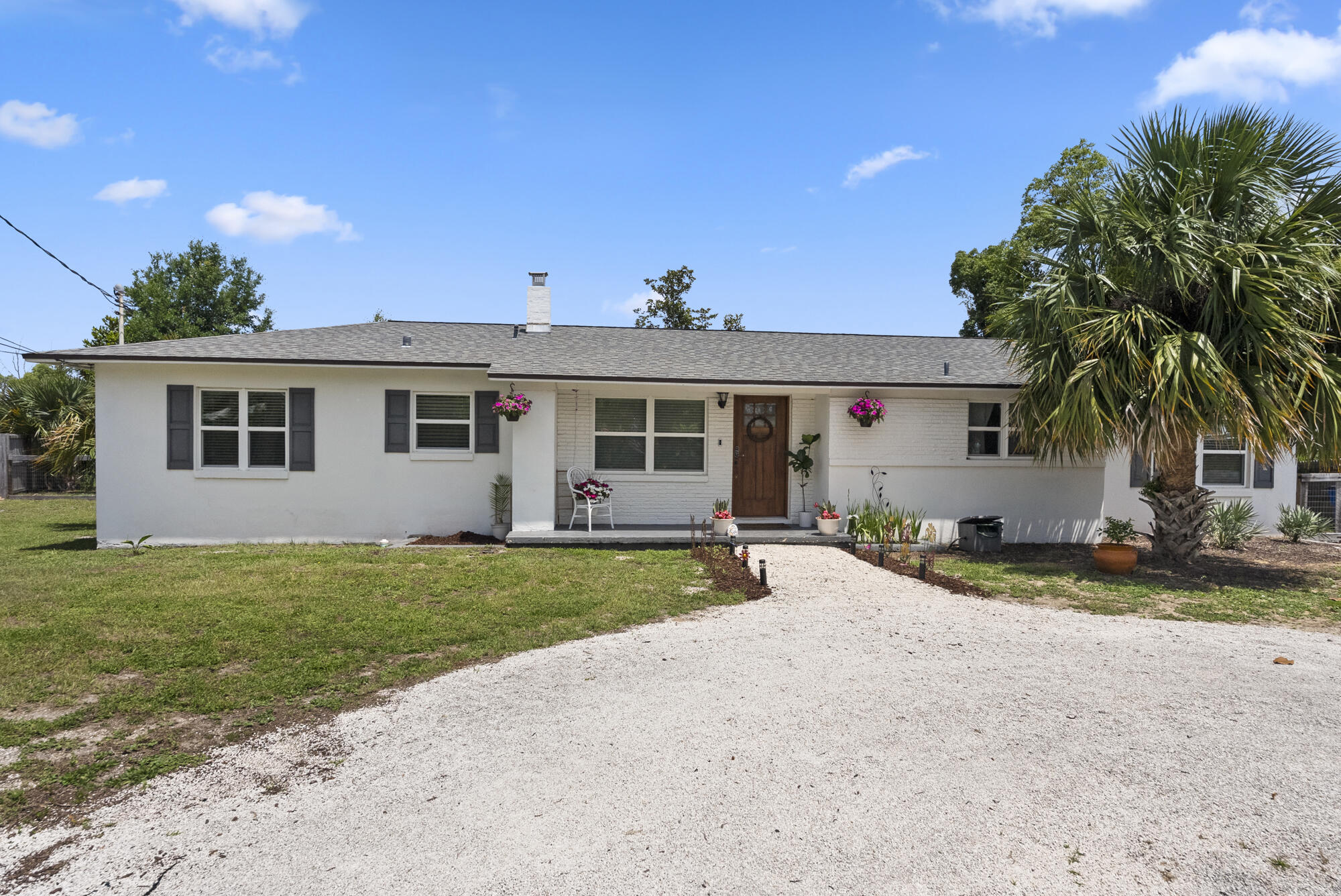 Single-story white house with a wooden front door, gravel driveway, and large palm tree.