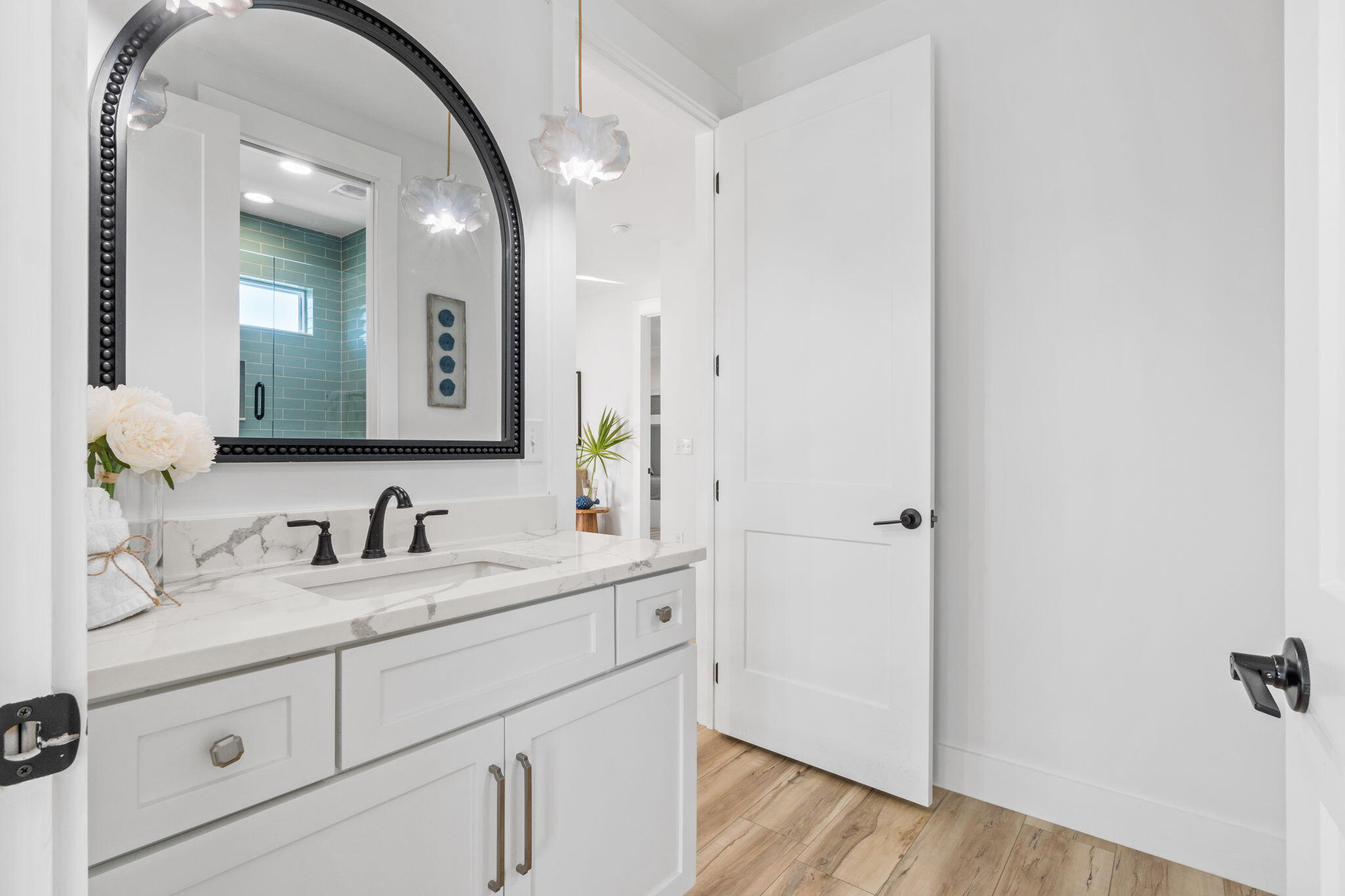 Bright bathroom with a white marble vanity, arched black mirror, and teal-tiled shower.