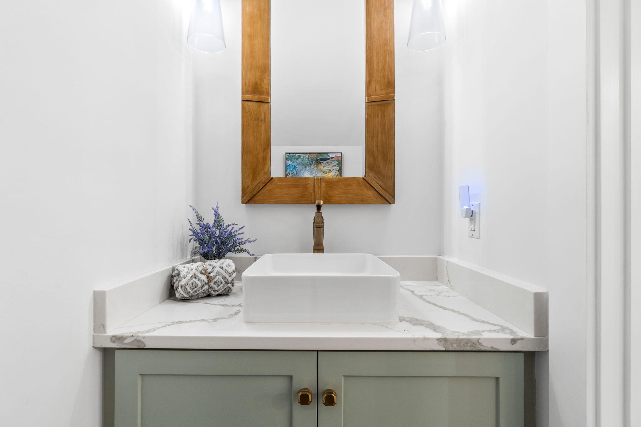Bathroom vanity with a white vessel sink, veined countertop, sage-green cabinets, and wood-framed mirror.