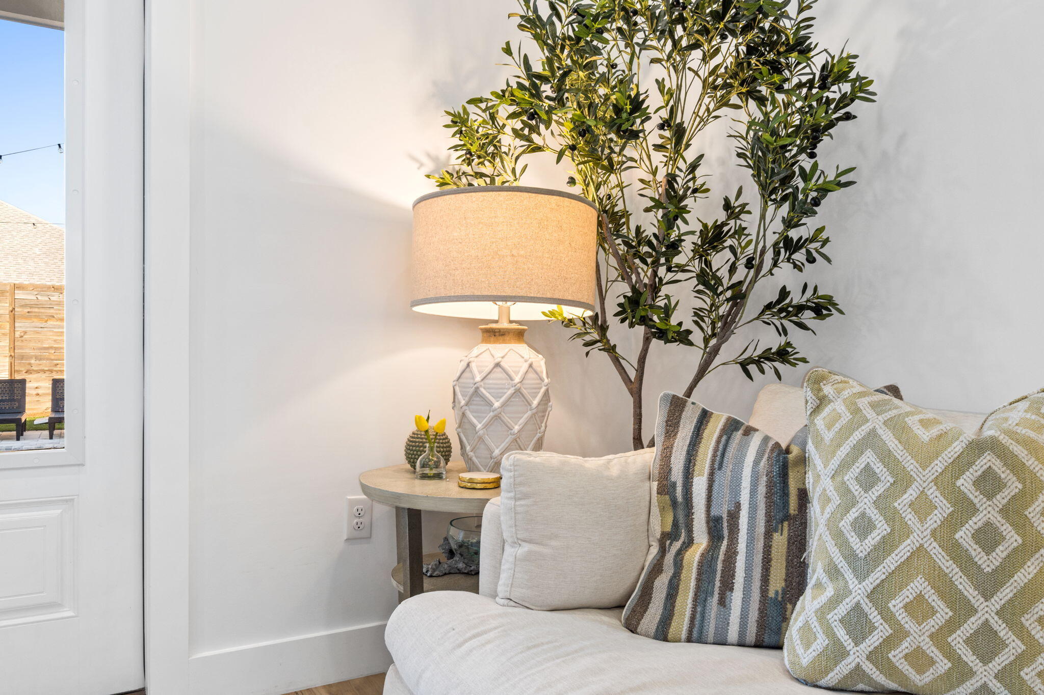 Living room sofa with patterned cushions beside a lit table lamp and leafy indoor tree.