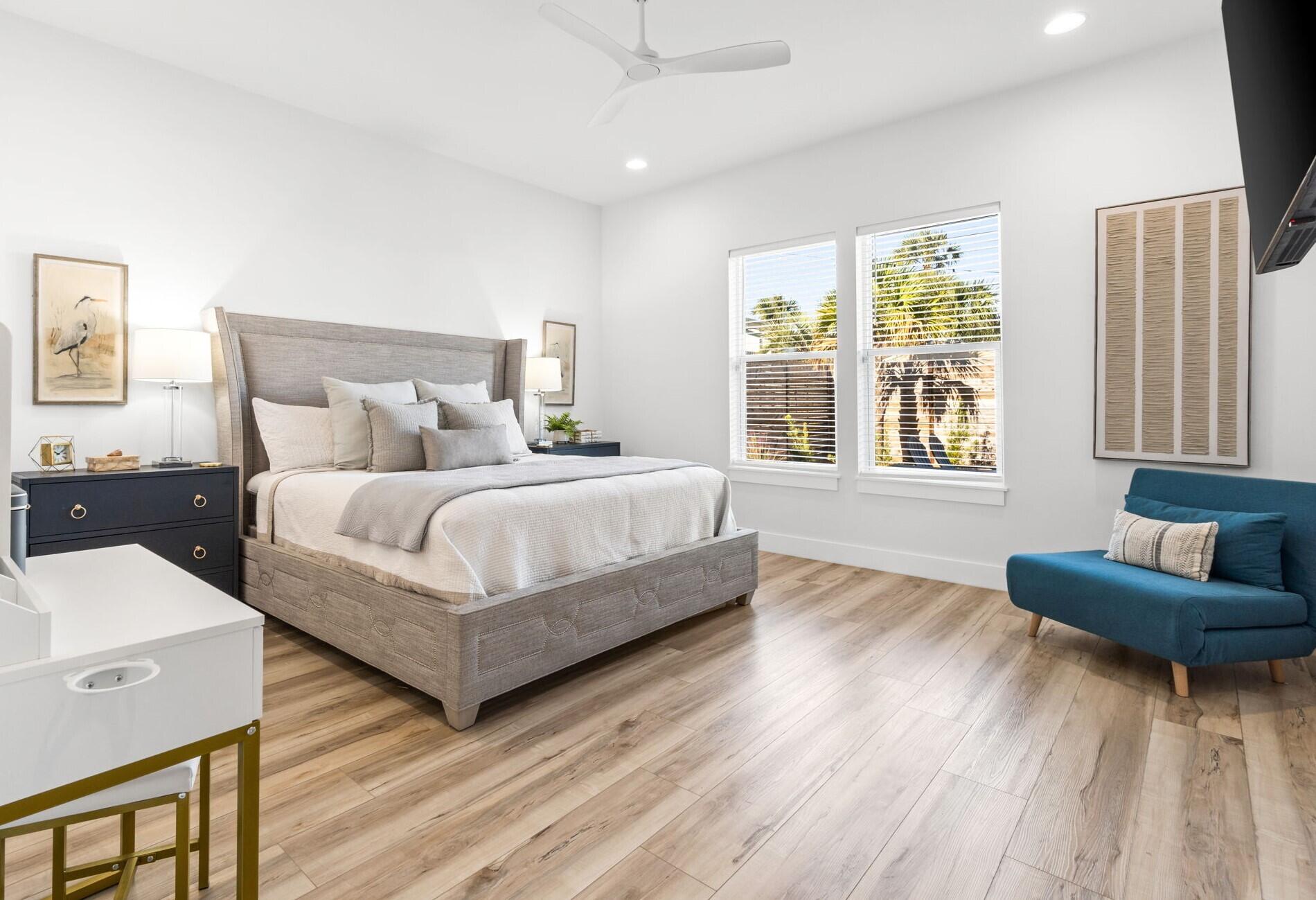 Bright bedroom with a large upholstered bed, teal loveseat, and windows overlooking palm trees.
