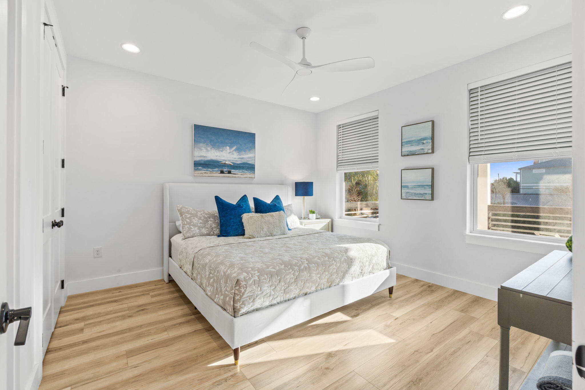 Bright bedroom with a white bed, blue accent pillows, light wood floors, and windows with blinds.