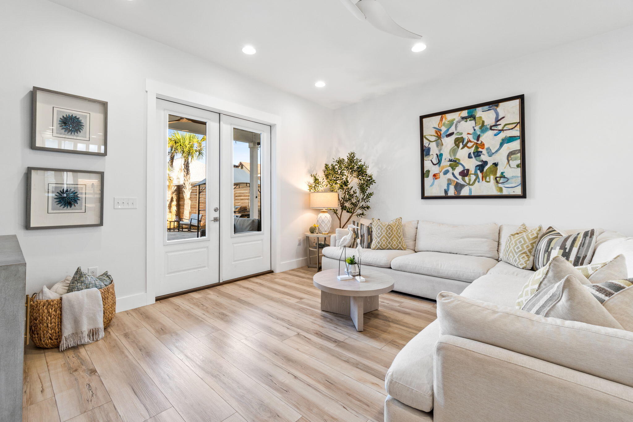 Staged living room with white sectional sofas, light wood floors, and glass doors to a patio.