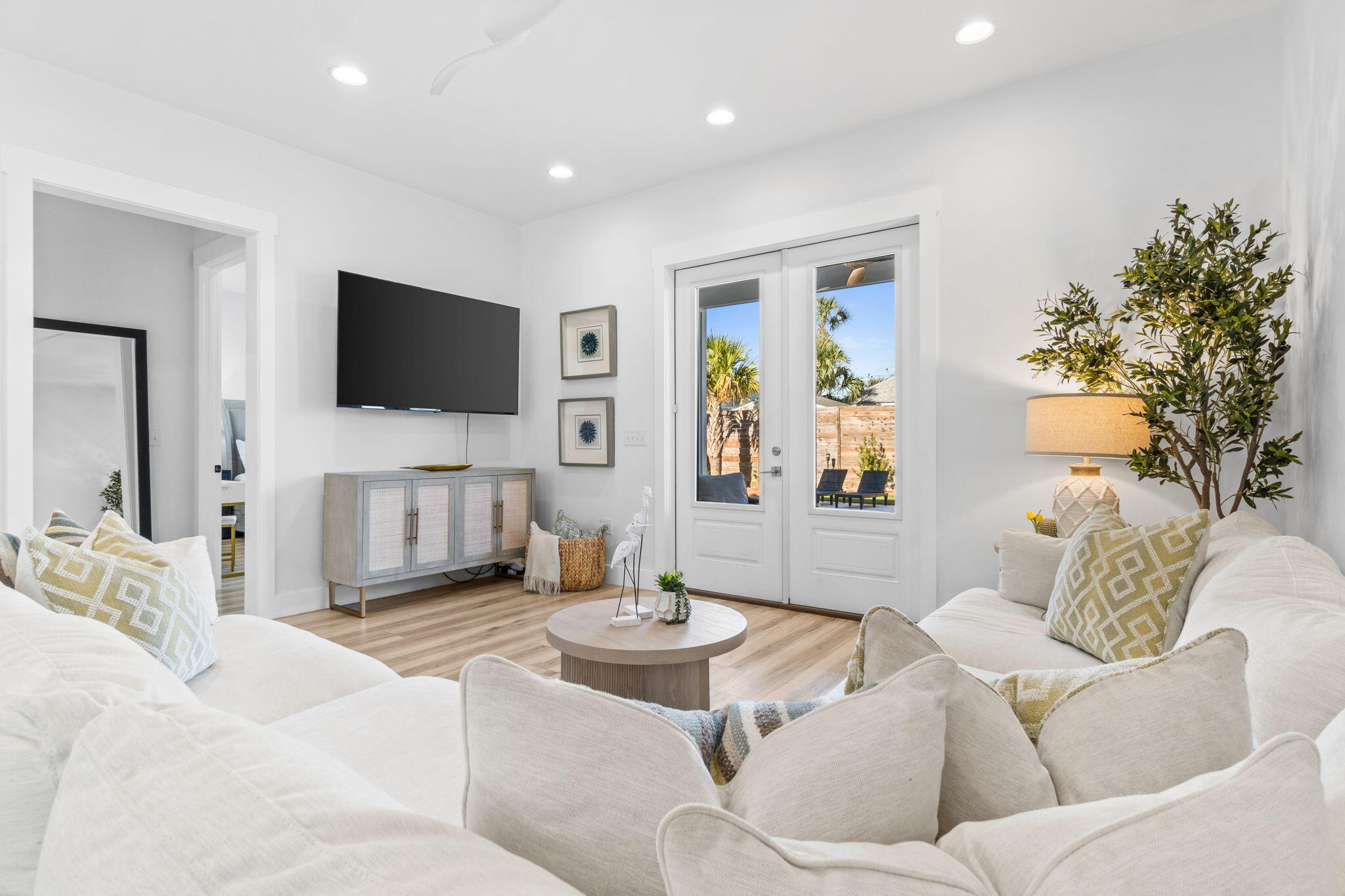 Bright living room with white sofas, a wall-mounted TV, and French doors to a palm-lined yard.