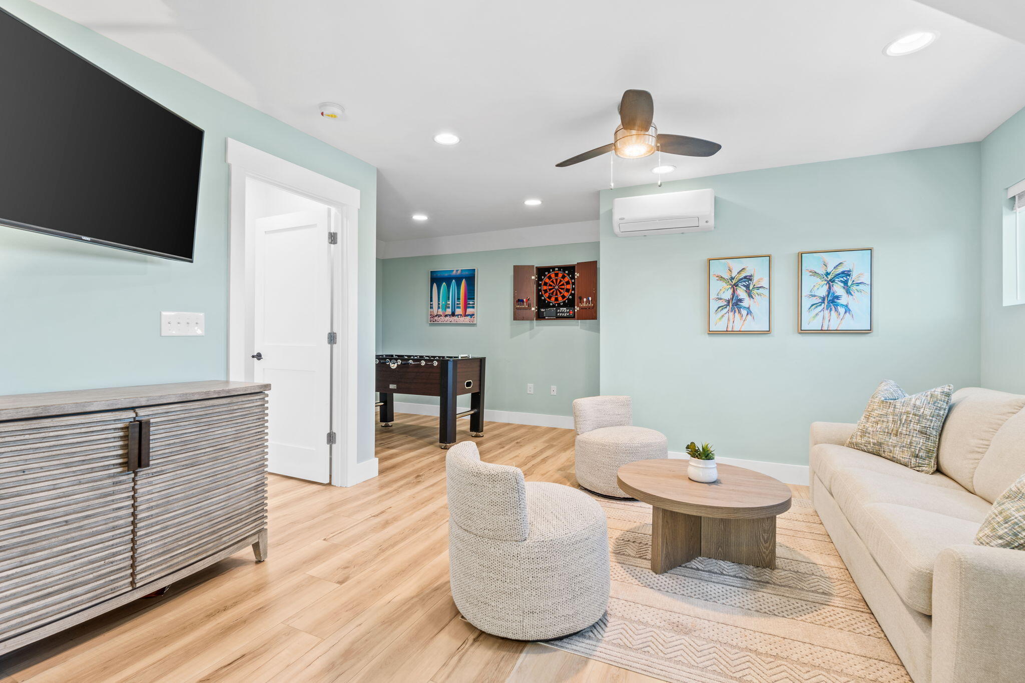 Coastal-style living room with a cream sofa, rounded chairs, and a foosball table beyond the doorway.