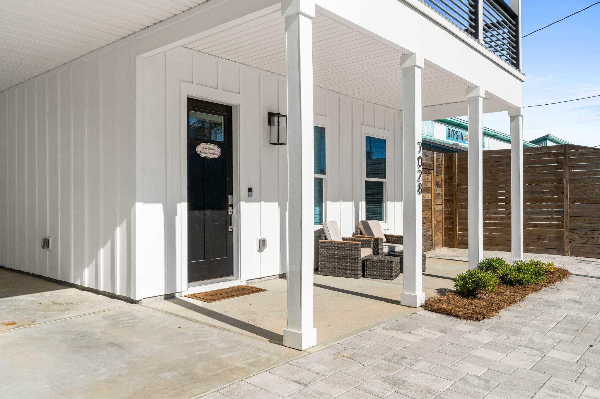 White home’s covered front porch with a black door, wicker seating, and wooden privacy fence.