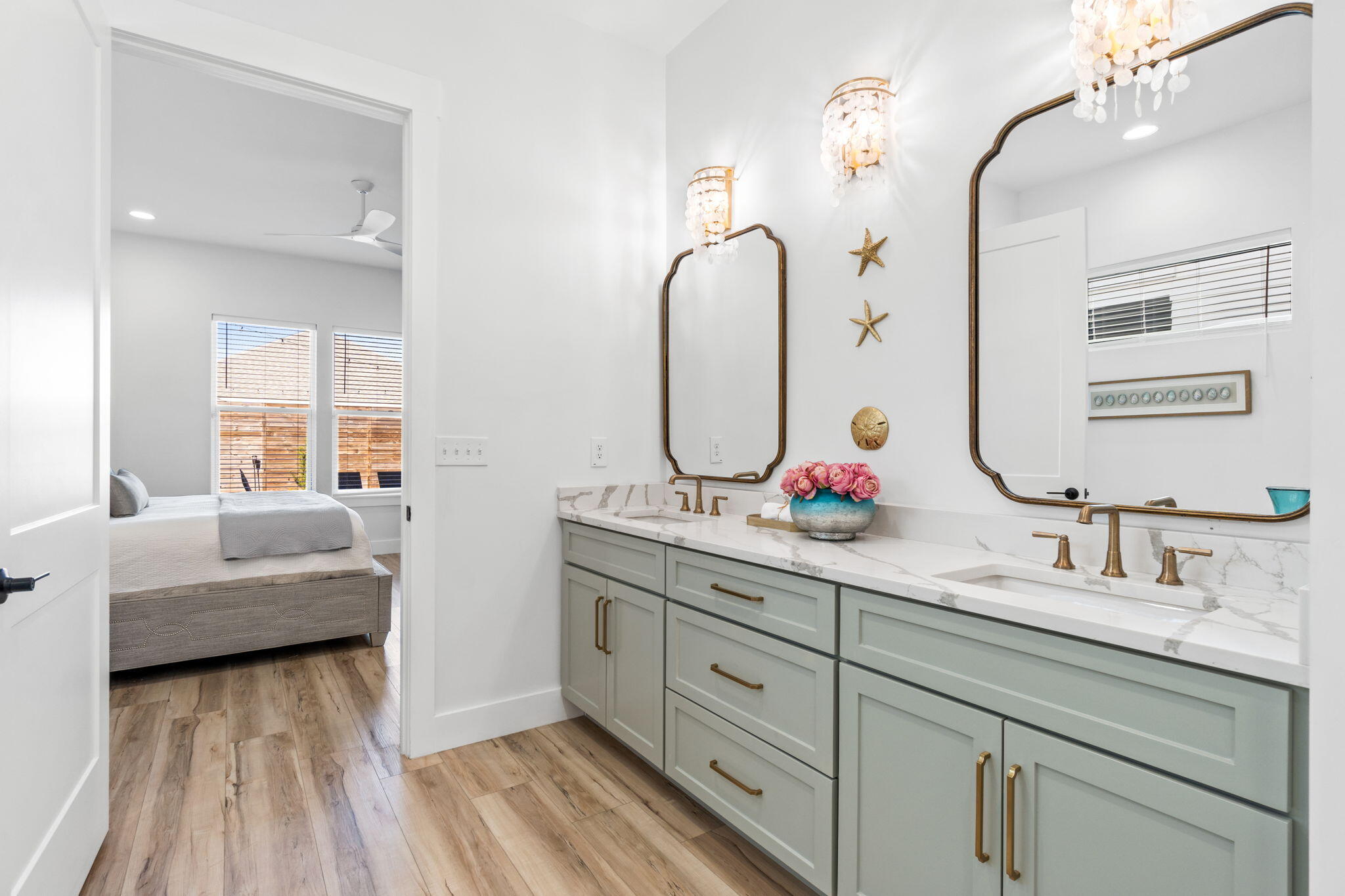 Bathroom with sage double vanity, marble-look counters, brass fixtures, and a bedroom visible through the doorway.